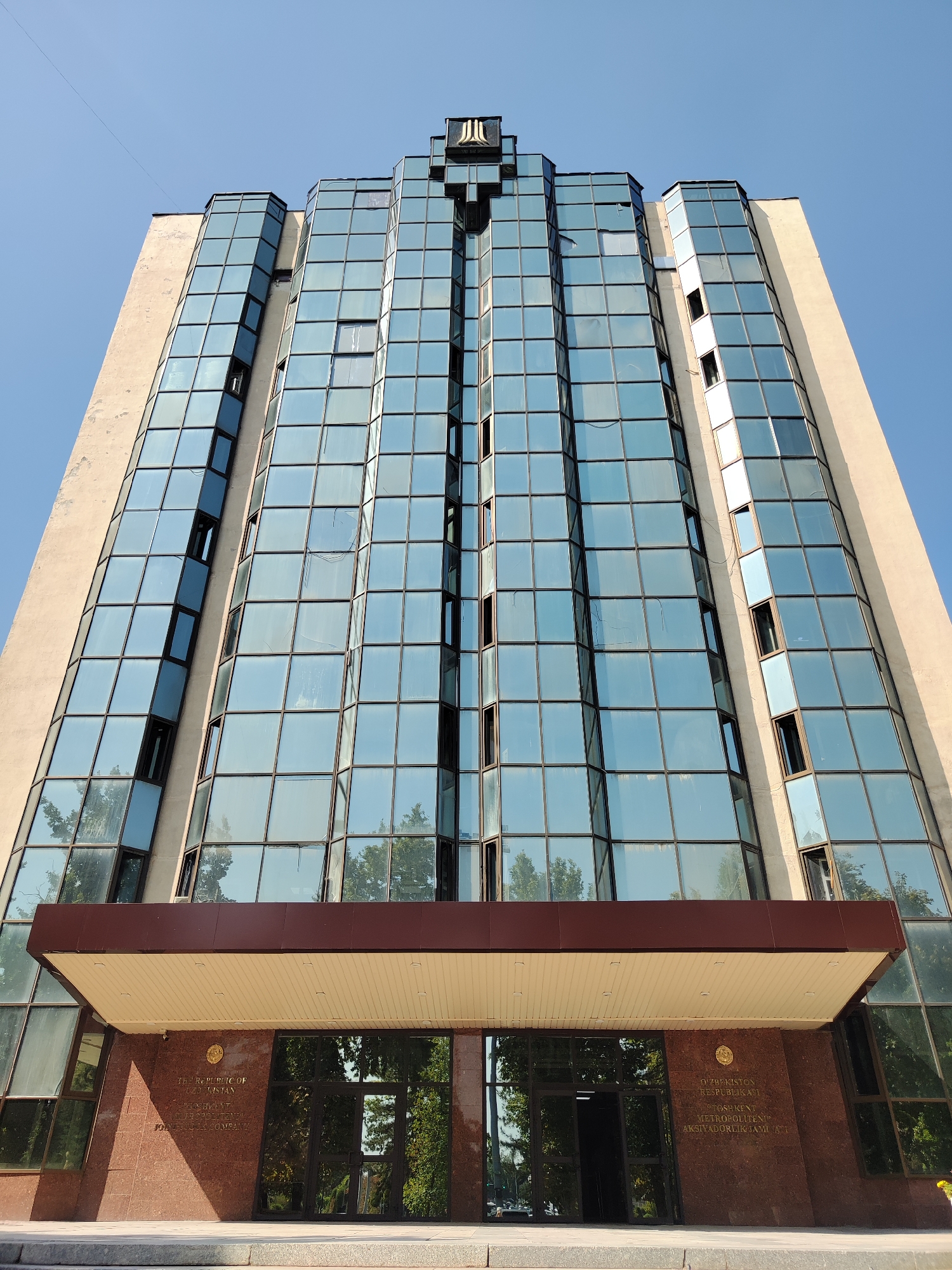 The image shows a low-angle, wide shot of the Tashkent City Administration Building (Hokimiyat) located in Tashkent, Uzbekistan. The building features a distinctive modern architectural style with a prominent glass facade that reflects the clear blue sky and surrounding trees. The facade is composed of numerous rectangular glass panes, some of which are open, revealing the interior. The building has a stepped, asymmetrical design, tapering towards the top. The lower section of the building is clad in reddish-brown granite, and features large glass doors leading into the building. Inscriptions in gold lettering are visible on the granite facade, indicating the building's official status. The entrance is covered by a dark brown canopy with a beige soffit. The scene is set against a bright blue sky, suggesting a sunny day. No people are visible in the image, and the focus is entirely on the architecture of the building.