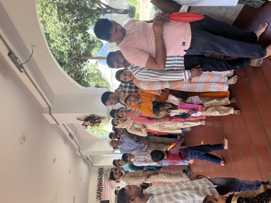 The image shows a queue of people waiting outside a building. It is a covered walkway with a white ceiling and columns. There are many people of different ages and genders in the queue. Some are wearing traditional Indian clothes, while others are wearing western attire. The people are standing in a line waiting for their turn. There is a small child in the line holding a pink toy, and a young man wearing a red vest is behind him. To the right of the queue, the walkway opens up to a view of green trees and white tent structures in the distance. The location of the image is a covered walkway outside a building in Panchkula, India, based on coordinates (latitude: 30.723, longitude: 76.862). The image is taken from a slightly high angle, looking down on the queue.