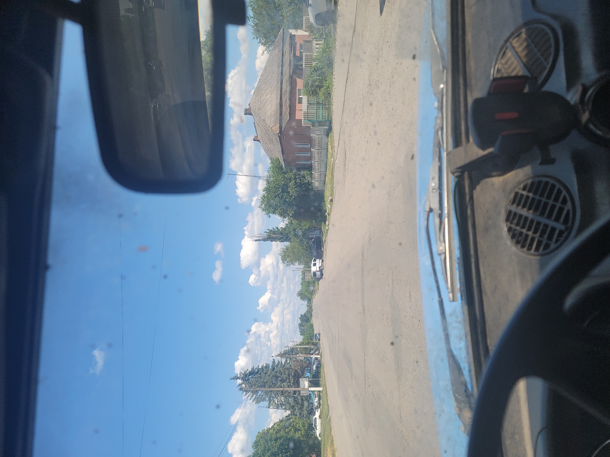 The image is a point-of-view shot from inside a car, looking out through the windshield at a street scene in Pereshchepyne, Ukraine. The sky is bright blue with scattered white clouds. The road ahead is asphalt, with parked cars and trees lining both sides. A residential house with a red facade and a gray roof is visible on the right side of the street, behind a white fence and green gate. The interior of the car is visible in the foreground, including the steering wheel and dashboard with a phone mount. The rearview mirror reflects a portion of the road and vehicles behind.