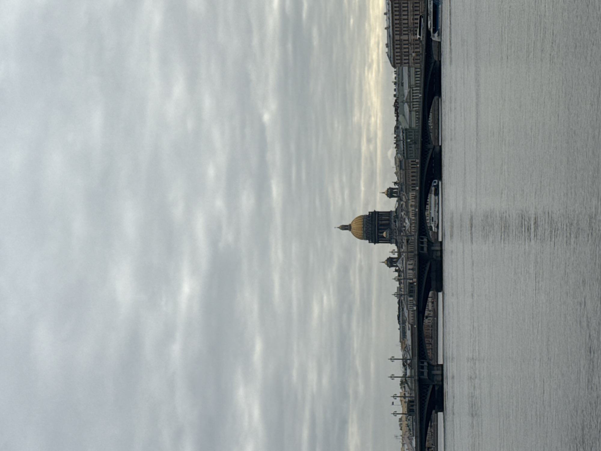 The image displays a wide, panoramic view of a city waterfront, observed under an overcast sky. A broad river, likely the Neva, occupies the foreground and mid-ground, its calm surface reflecting the muted grey light from above.
Along the far bank, a distinctive city skyline stretches horizontally. It features numerous multi-story buildings with classical and neoclassical architectural styles, characterized by their regular windows and sturdy construction. A prominent landmark on the left side of the skyline is a large, majestic golden dome, belonging to St. Isaac's Cathedral, which gleams subtly against the pale, cloudy backdrop.
Several elegantly arched bridges traverse the river, connecting the banks. These bridges are dark in color and appear to be adorned with classical streetlights along their length. Small boats or vessels are faintly visible moored near the riverbanks, particularly beneath the bridges.
The upper half of the image is filled with an expansive, uniformly overcast sky. The clouds exhibit soft layers of grey and white, suggesting a calm and subdued daytime atmosphere. No individuals are visible in the scene. The overall mood is serene and somewhat quiet, characterized by the consistent grey tones of the sky and water. The architectural cues, especially St. Isaac's Cathedral, firmly place the location in Saint Petersburg, Russia. There is no visible text.