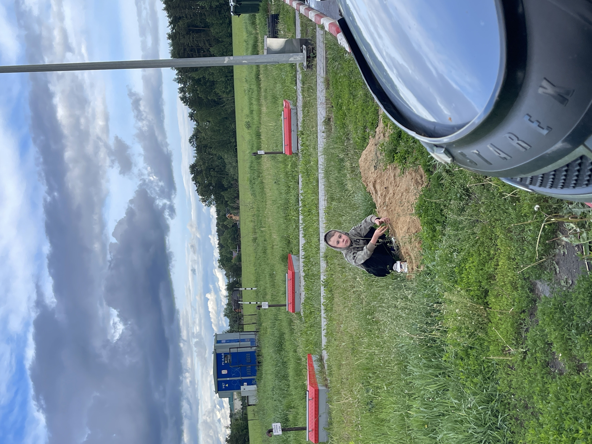 A photo taken outdoors in Бавыкино, Russia, shows a boy crouching next to a dirt pile with a green grassy field behind him. Behind the boy, several red and gray markers stand in the field. Further back, a blue metal structure, identified as equipment building ПУ-Р-4х4В, is situated behind a chain-link fence. In the top right, there's part of a black object with the word "WERK" visible. The sky above is filled with cloudy formations.