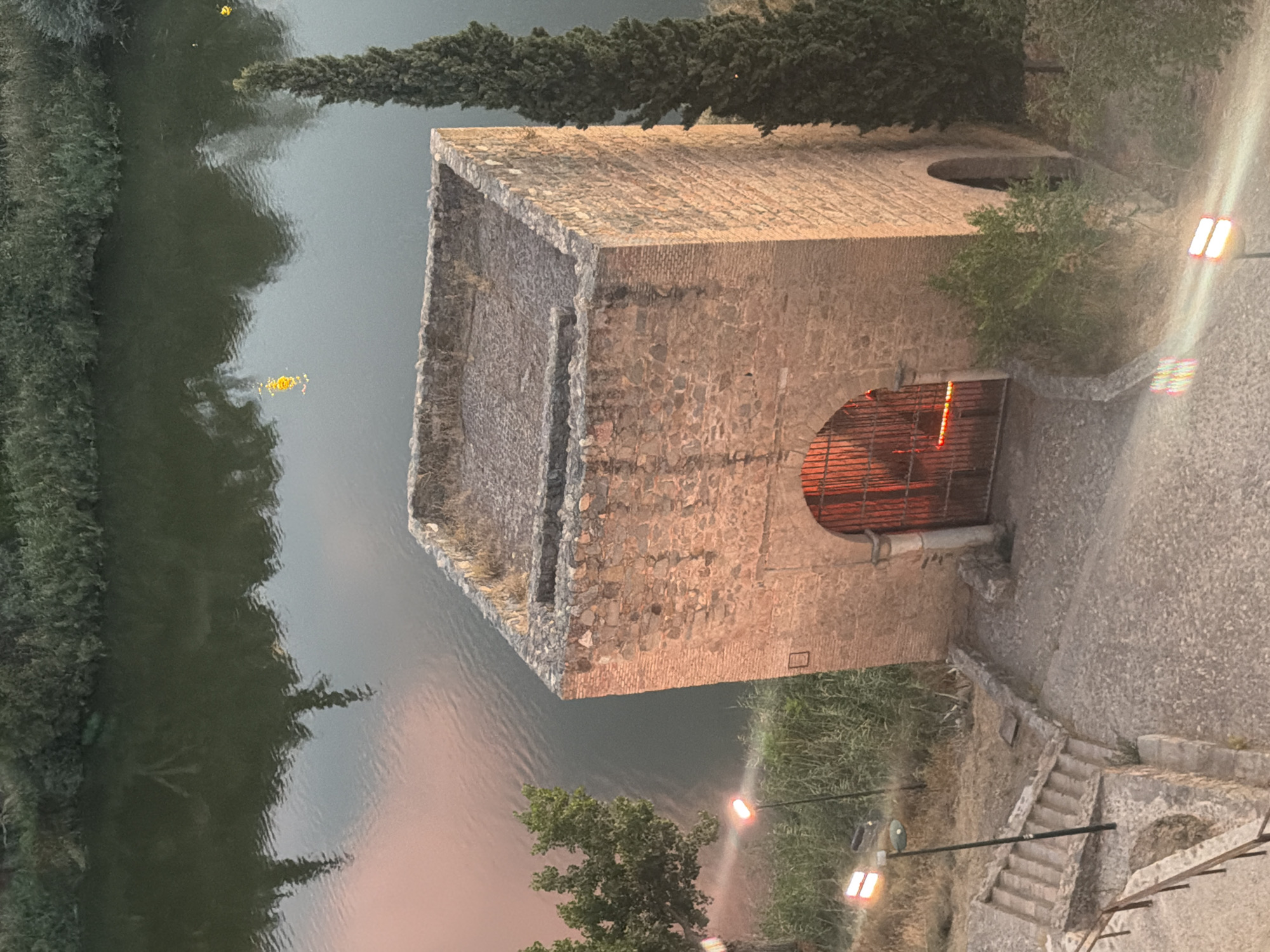 This is an aerial, high-angle photograph of a medieval stone tower situated beside a river in Toledo, Spain. The tower is roughly square in shape, with weathered stone walls and a flat, gravel-covered roof. An arched entrance at the base of the tower is sealed with a dark, ornate metal gate, through which a red glow emanates. The river, appearing dark and reflective, flows alongside the tower, with lush green vegetation lining its banks. In the foreground, a cobblestone pathway winds up towards the tower, interrupted by a series of stone stairs and lit by modern street lamps. A tall, slender evergreen tree stands to the right of the tower. The overall scene suggests a historical setting bathed in the soft, warm light of either dawn or dusk.