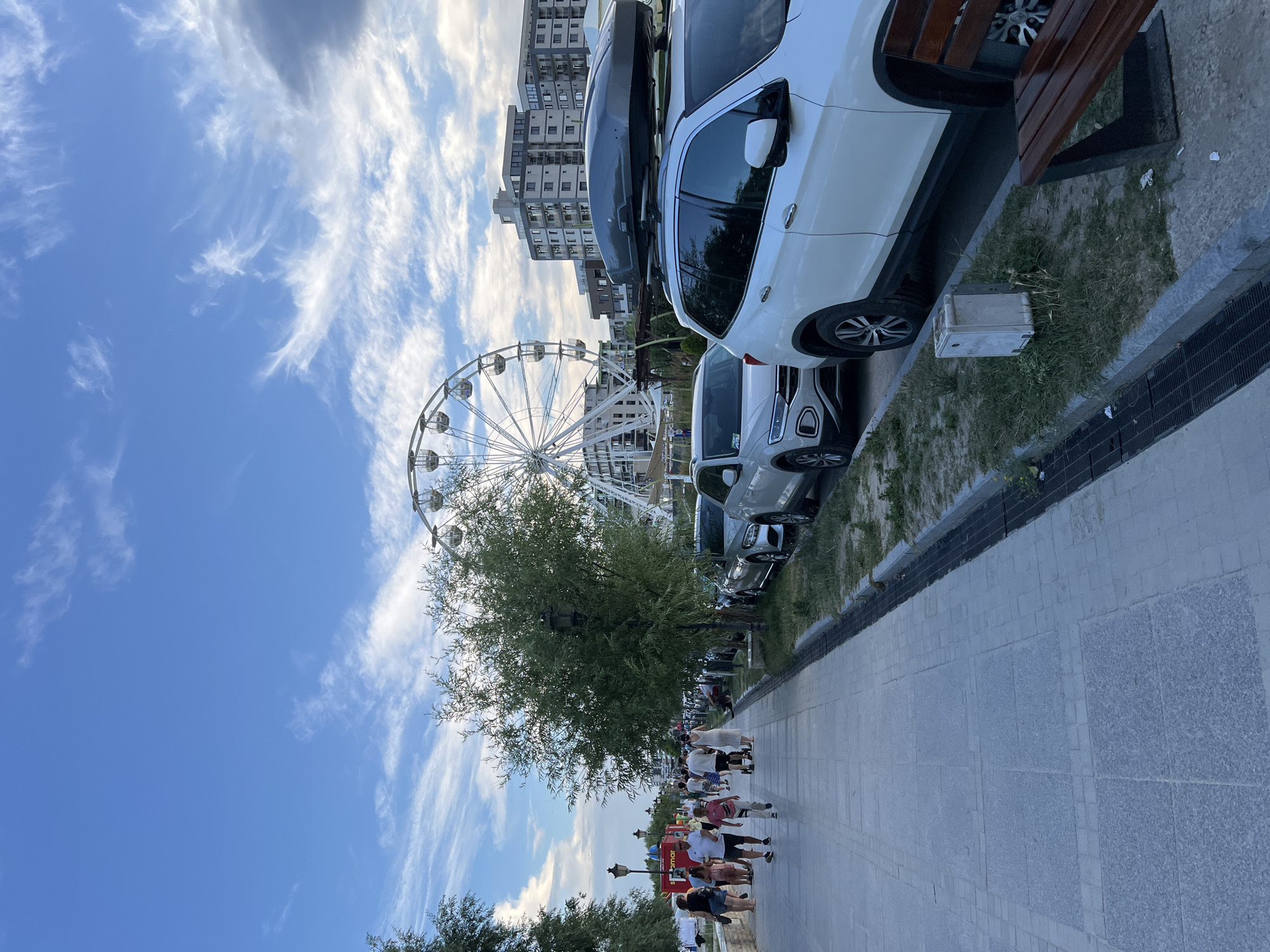 The image depicts a bustling promenade in Năvodari, Romania, with a Ferris wheel and modern buildings visible in the background. A paved walkway runs along the left side of the image, populated by numerous pedestrians strolling and interacting. To the right of the walkway, several parked cars are lined up, some equipped with roof carriers. A large green tree stands between the walkway and the parked cars, with streetlights visible intermittently. The sky is partly cloudy, with a mix of blue sky and wispy white clouds. A wooden bench is positioned on the right side of the image, overlooking the promenade. The overall scene suggests a lively recreational area, likely a popular spot for tourists and locals.