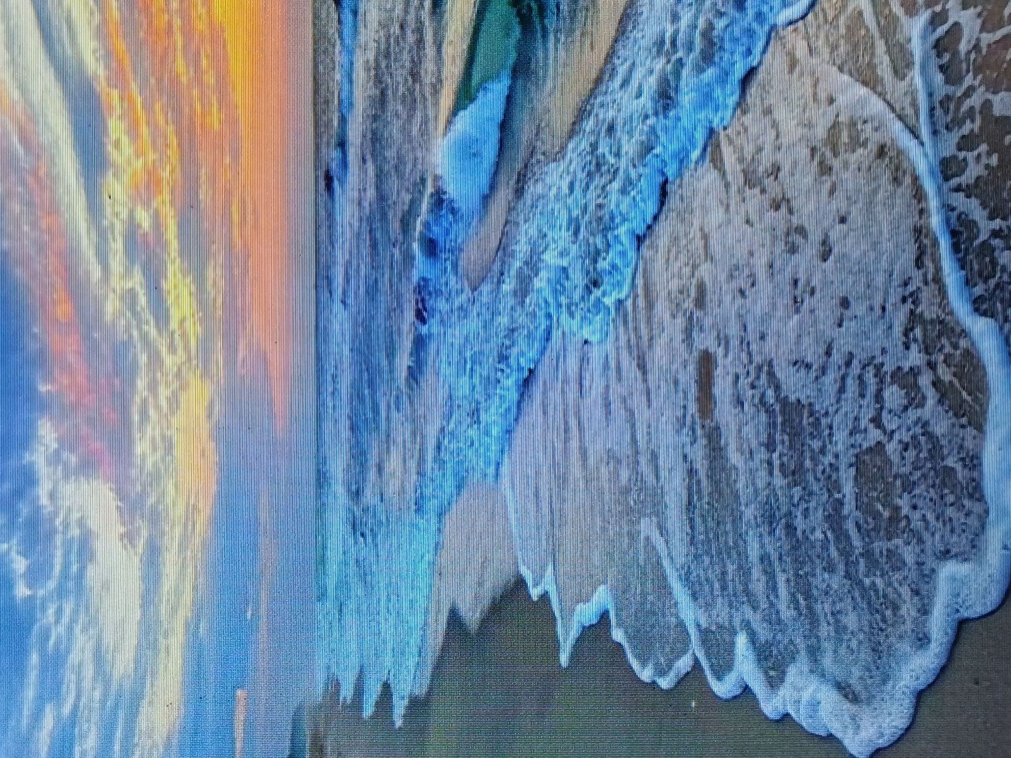 The image shows a beach scene at sunset. On the left, the sky is a mix of orange, yellow, and blue colors, indicative of the setting sun. The right side of the image features a wave crashing onto the shore, with white foam and turquoise water dominating the scene. The sand is visible beneath the water, showing a mix of wet and dry areas. The location is likely in or near Juan Griego, Venezuela, as that was specified in the prompt.