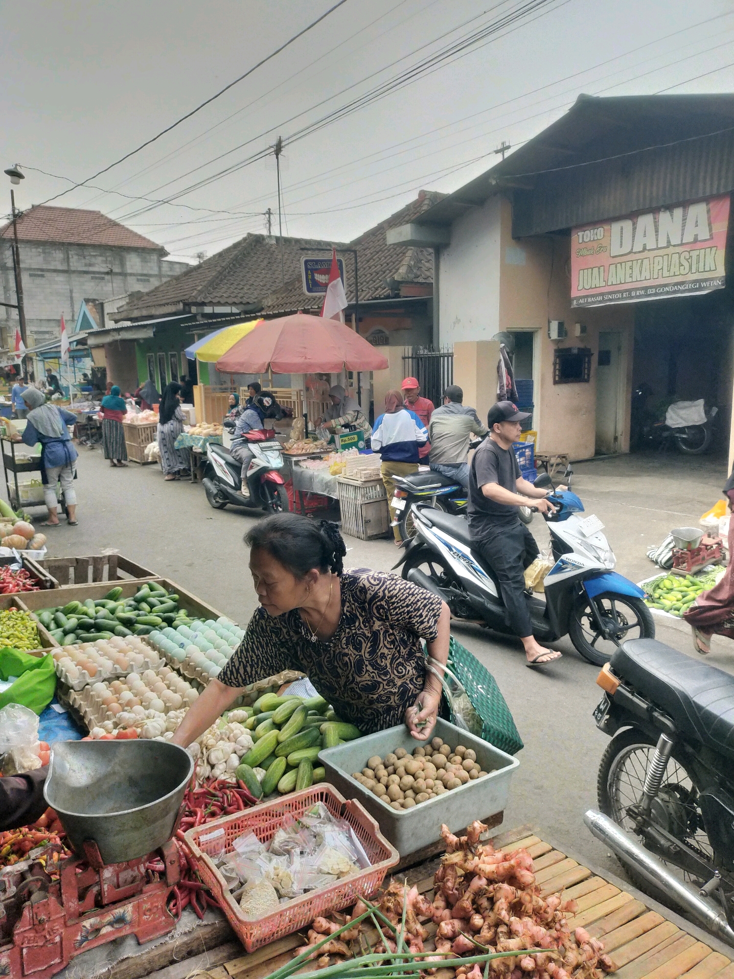 The image depicts a street market scene in Gondanglegi, Indonesia, with a focus on a woman vendor in the foreground. She is wearing a dark, patterned top and is attending to a display of produce. In front of her are several crates and baskets filled with various goods, including red chilies, garlic, cucumbers, eggs, and small brown fruits or vegetables. A large metal scale sits on a wooden surface, partially obscuring the chili display. To her right, there is a pink plastic basket containing small, sealed plastic bags of what appear to be spices or seasonings. Further back, in the middle ground, other vendors and customers are visible. A man in a black t-shirt and cap is riding a blue and white motorcycle. Another motorcycle is parked behind him. Umbrellas, some red and yellow, are providing shade over some of the stalls. In the background, buildings with tiled roofs line the street, and a banner that reads "TOKO DANA JUAL ANEKA PLASTIK" is visible on one of the structures, indicating a plastic goods store. The environment appears to be an outdoor market on a street with overhead power lines and a cloudy sky.