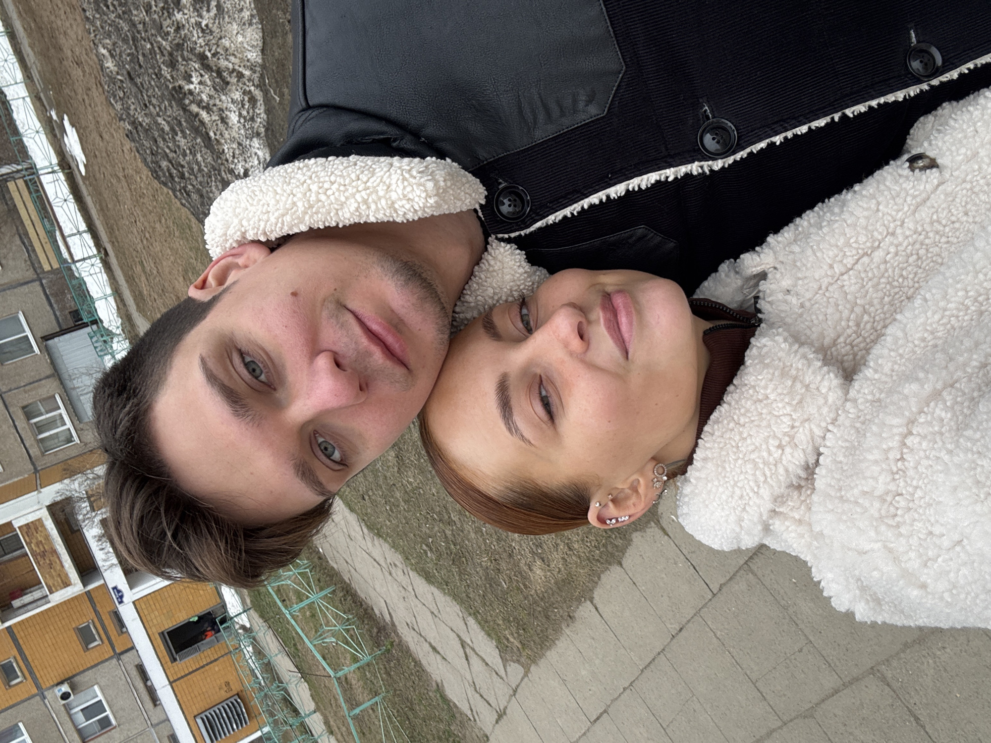A selfie-style, close-up shot captures a man and a woman lying on what appears to be a concrete surface, likely a sidewalk or pavement. The man, on the left, has short brown hair, fair skin, and green eyes. He is wearing a black jacket with a white shearling collar. The woman, on the right, has fair skin, auburn hair slicked back, and multiple earrings in her left ear. She is wearing a white shearling jacket. Both are looking towards the camera. The background features a cityscape, including multi-story apartment buildings with peeling paint, visible through green fences, and patches of dead grass. Based on the building type and surrounding details, this is possibly in the city of Chaikovsky, Russia.