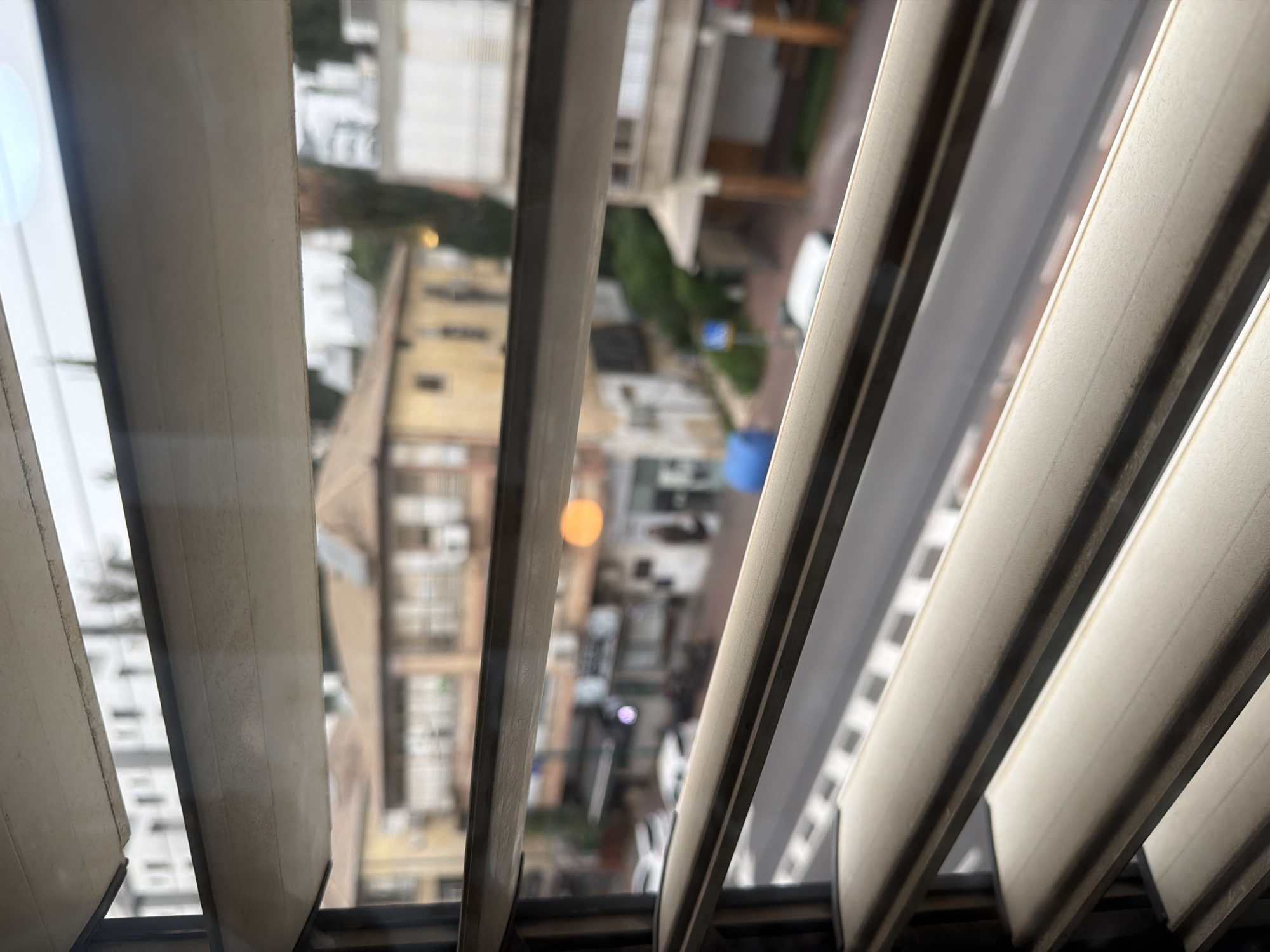 View from behind closed blinds in Ramat Gan, Israel. Outside the window, a street with buildings and cars is visible, though the view is obscured by the blinds. There is a blue trash can and an orange street light.