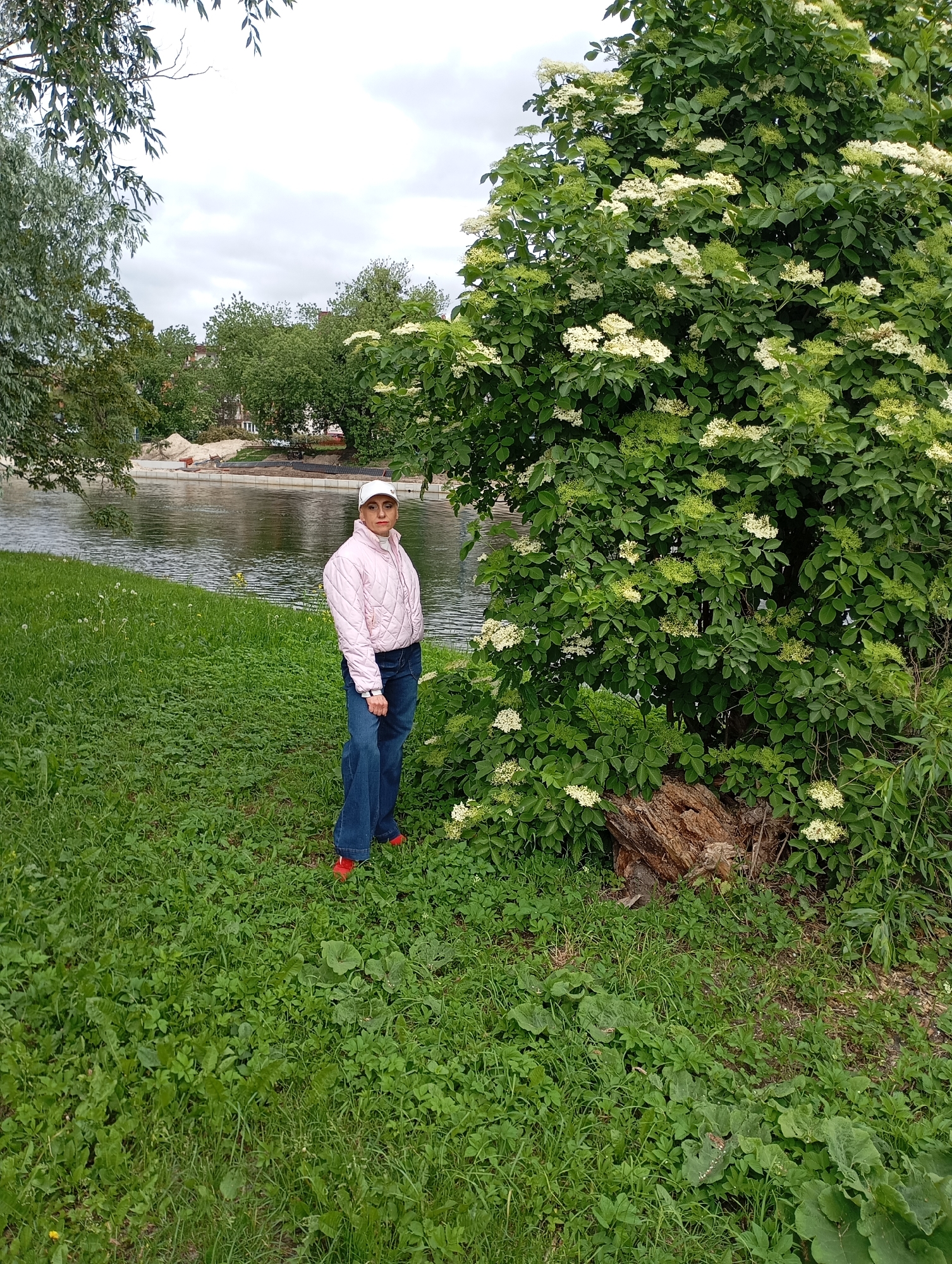 The image captures a woman standing next to a flowering tree on a grassy bank along the Brda River in Bydgoszcz, Poland. The woman wears a white baseball cap, a light pink quilted jacket, blue jeans, and red shoes. She is positioned in the foreground, adjacent to a large tree blooming with white flowers. Behind her is the river, reflecting the overcast sky and surrounding greenery. Across the river, a construction site is visible along the bank, with exposed soil and equipment. Further back, other trees and buildings form the cityscape. The grassy bank is covered in lush, green vegetation.