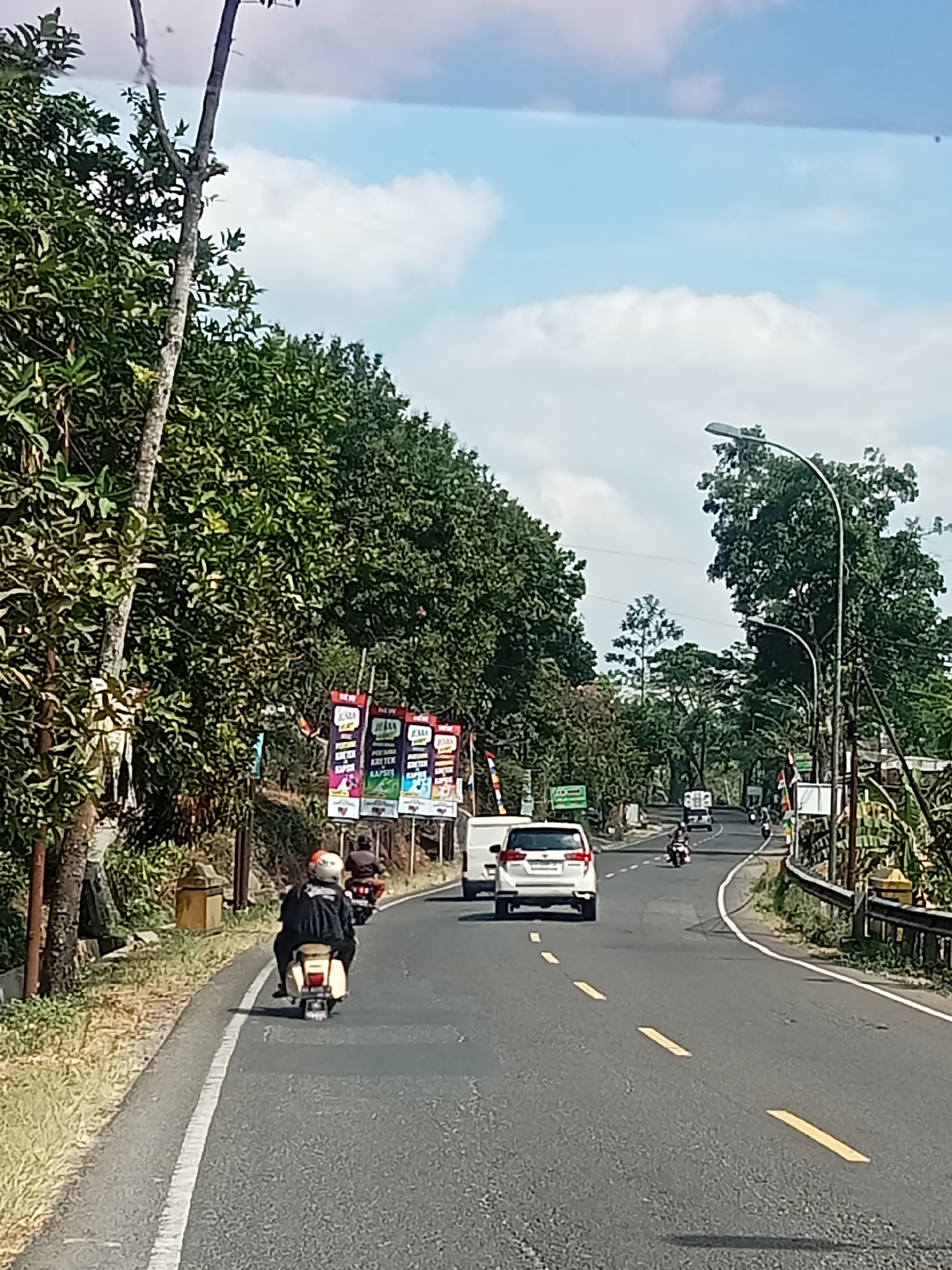 The image depicts a roadside scene in Selokromo, Indonesia, on a sunny day. A two-lane asphalt road curves gently to the right, bordered by lush green trees on both sides. A white car is traveling away from the camera in the right lane, while several motorcycles are also on the road. In the foreground, a person is riding a beige scooter in the left lane, facing away from the camera. They are wearing a black jacket and a white and orange helmet. The sky is blue with scattered white clouds. Along the left side of the road, a narrow grassy shoulder is visible, with a simple stone structure. On the right side, there is a metal guardrail. Several vertical banners with colorful graphics are displayed along the roadside in the background. The overall impression is of a rural or semi-rural road in a tropical or subtropical environment.