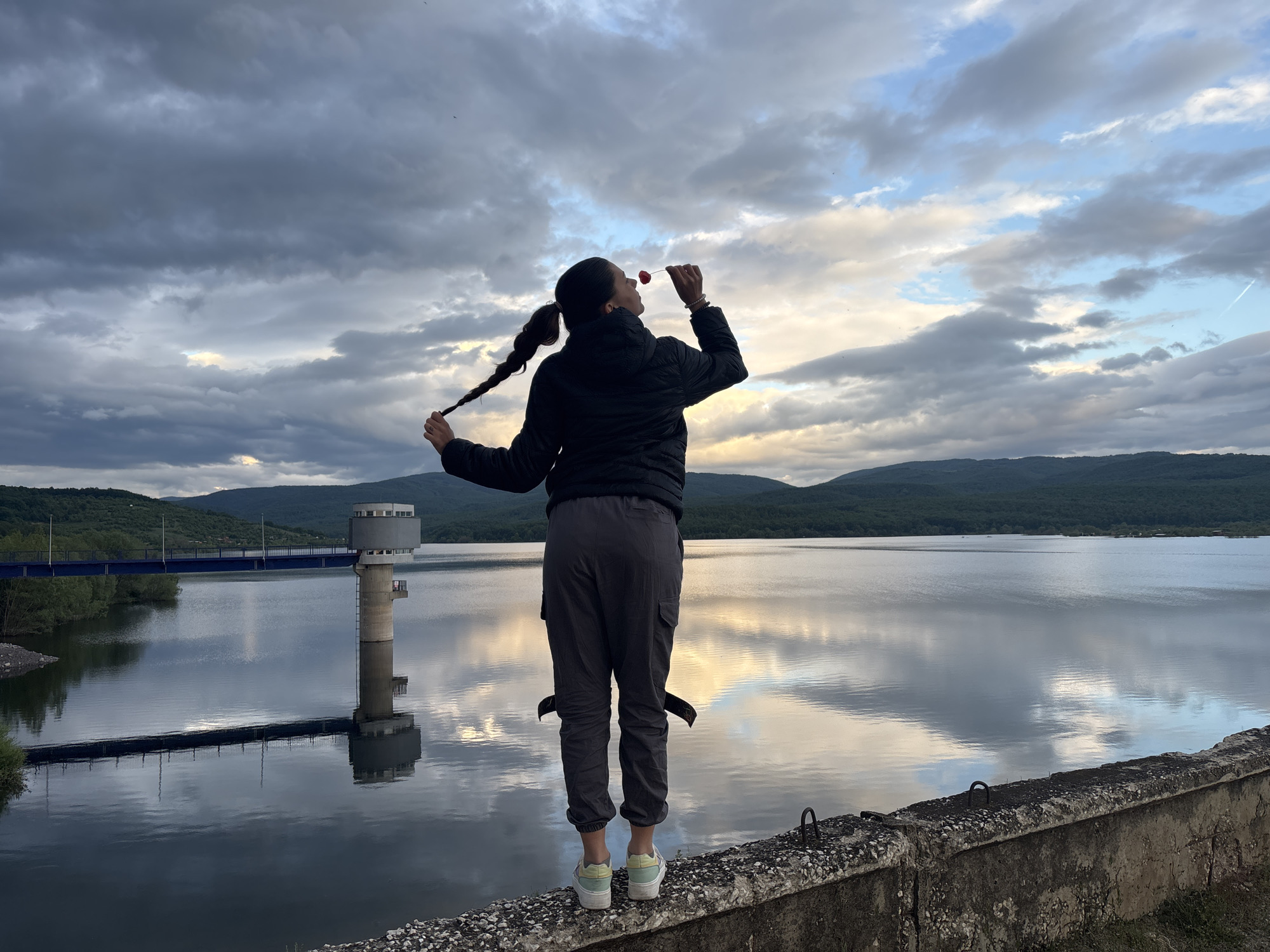 A young woman stands on a concrete ledge overlooking a large body of water with a tower and bridge in the background, likely the Ognyanovo reservoir in Bulgaria. She has her back to the viewer, holds a red fruit (possibly a cherry) up to her mouth, and holds one of her braids in the other hand. She wears a black jacket, grey pants, and light-colored sneakers. The sky is cloudy with patches of blue and orange visible. The water reflects the sky and surrounding landscape.