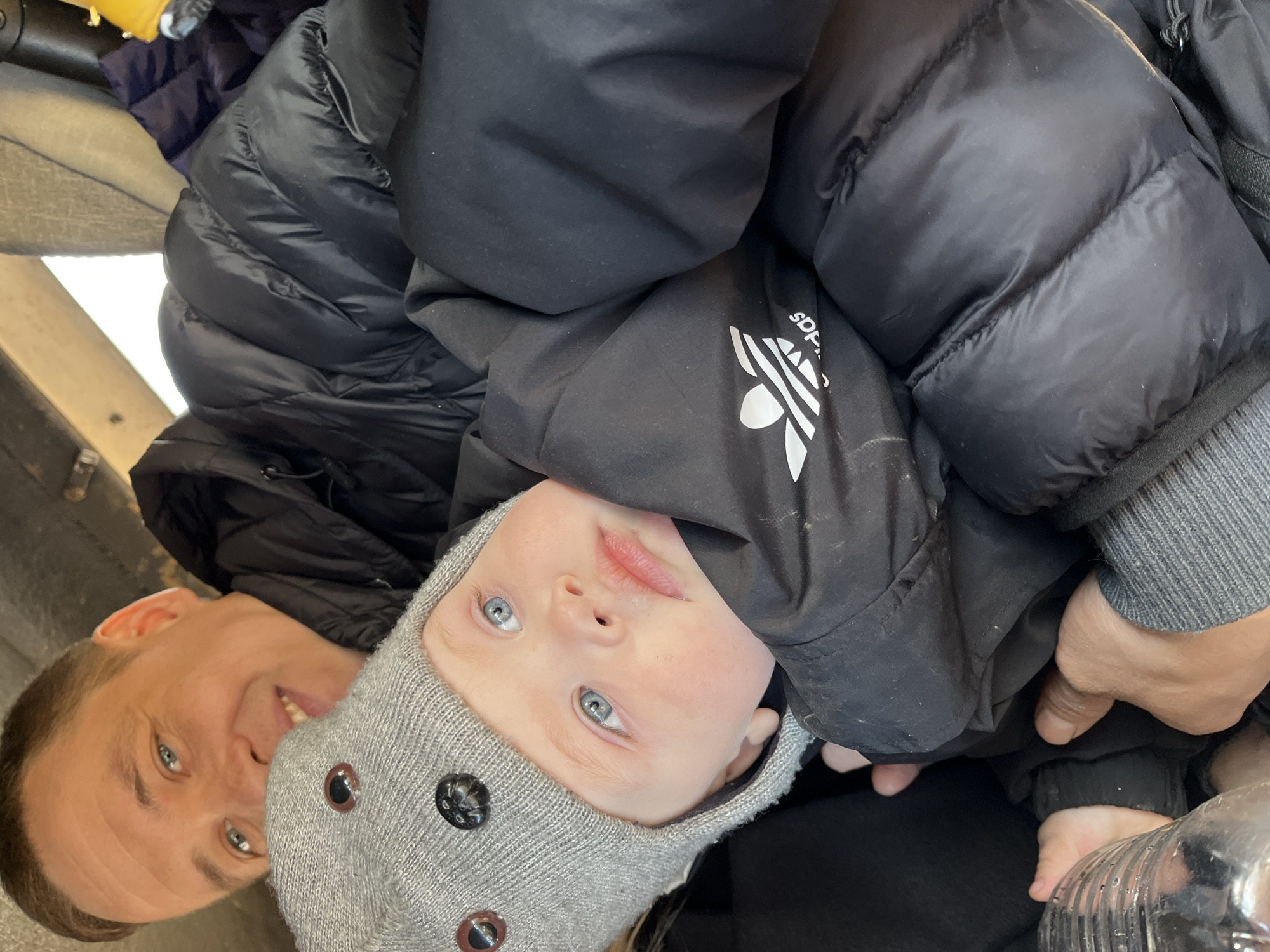 A medium shot shows an adult and a baby in an indoor location. The image appears to be taken inside a vehicle. The adult, with short brown hair, smiles at the camera. The baby, wearing a gray knit cap with decorative buttons, looks directly at the camera with blue eyes. Both are bundled in dark winter jackets. The baby's jacket appears to have an Adidas logo. A water bottle is visible near the bottom right corner of the frame. The location is identified as Chaikovsky, Russia.