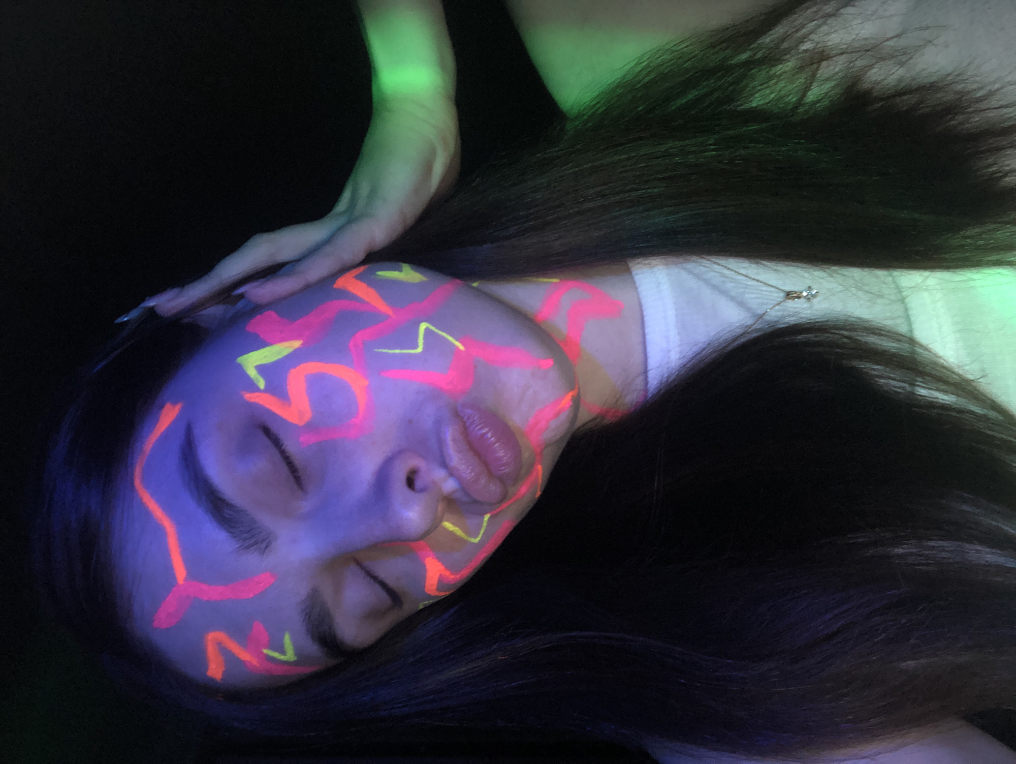An individual is captured in a close-up, artistic shot, showcasing face paint under blacklight conditions, likely in Noviy, Russia. The subject, a person with long, dark hair, is shown with their eyes closed and lips slightly parted, conveying a serene or contemplative expression. Their face is extensively covered in abstract, glowing patterns of bright pink/orange and yellow-green paint, which fluoresce vibrantly under ultraviolet light. The hair, skin, and a visible hand and forearm also exhibit a subtle purple-blue and green glow from the ambient lighting. A thin necklace with a small, clear pendant hangs around the neck, resting on a white garment, possibly a t-shirt. The background is deep black, emphasizing the luminosity of the paint and the subject. The scene suggests a themed event, performance, or artistic photography session utilizing blacklight effects.