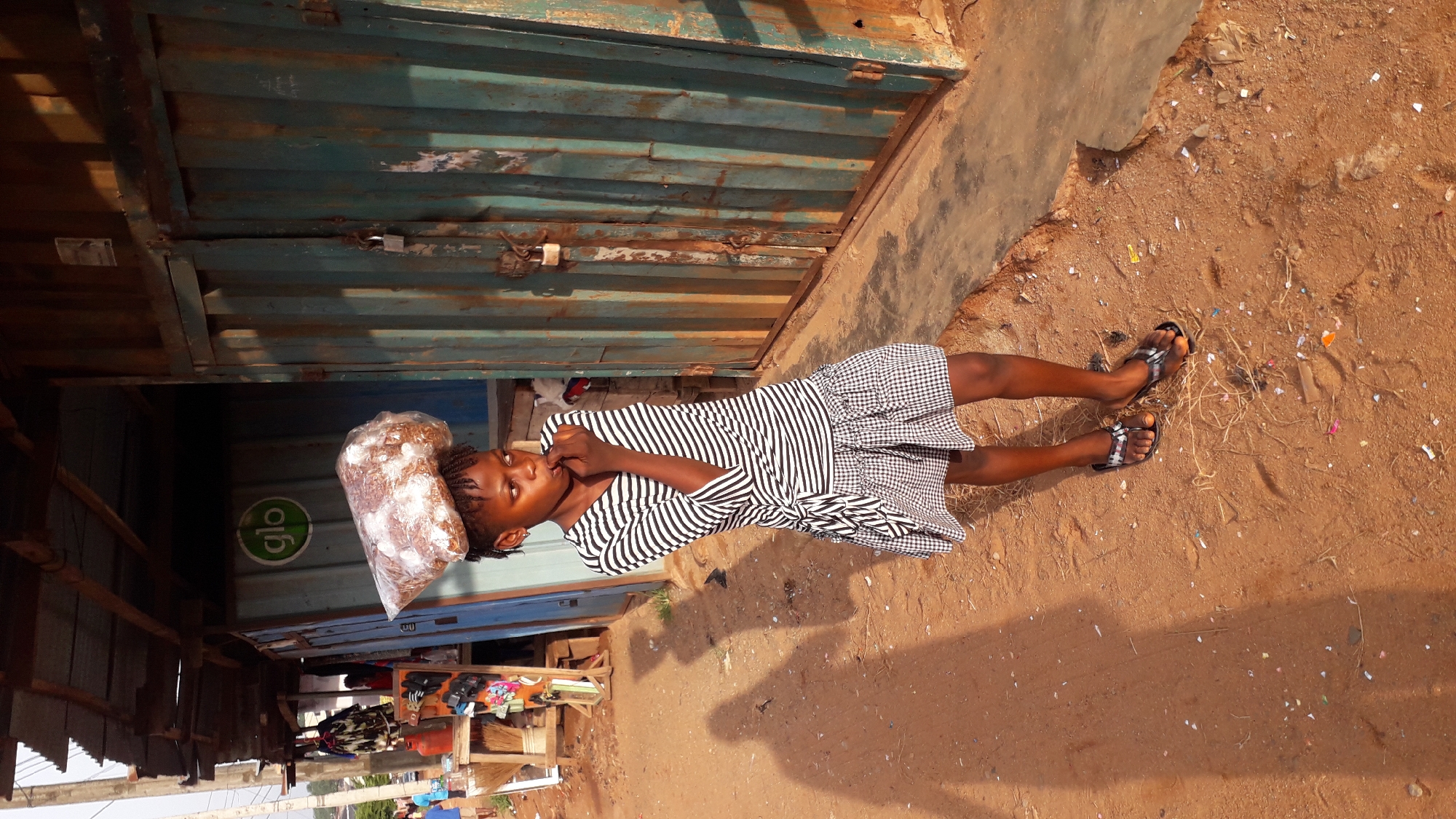 A young person stands in the sandy soil beside a building in Nkpor, Nigeria. The person, wearing a striped top and a skirt, is touching their face and holding a large plastic bag of an indiscernible product. The building behind them is made of corrugated metal and appears to be a shipping container converted into a retail space. The container has a green "Glo" logo on it. The ground is covered in trash.