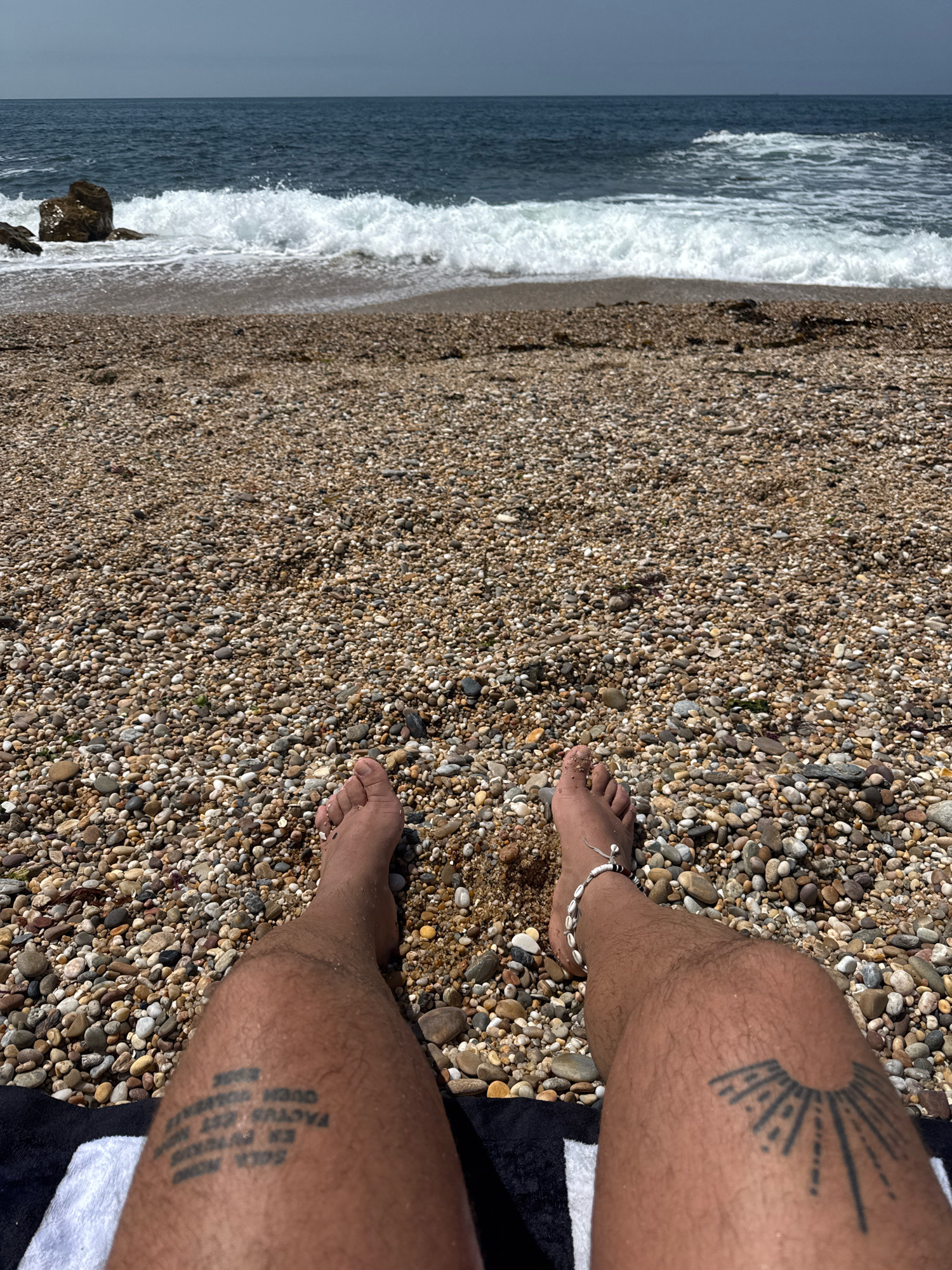 The image shows a first-person perspective of a person sitting on a pebble beach, with their legs and feet in the foreground. The beach is covered in small, varied-colored pebbles. The ocean is visible in the background, with waves crashing on the shore. The sky is overcast. There are tattoos on both legs: a circular, radiating sunburst on the right thigh and text on the left calf. The person is wearing a shell anklet on their right ankle. The scene is located on the coastline of Vila Nova de Gaia, Portugal.