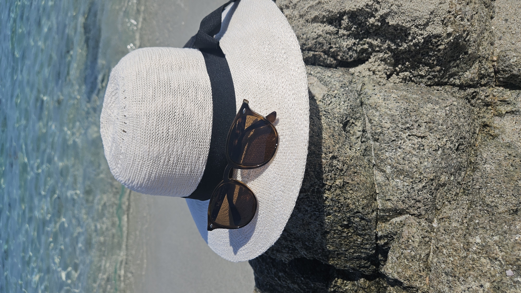 A wide-brimmed white hat with a black ribbon is resting on a textured rock at the edge of a sandy beach. Brown-tinted sunglasses are placed on top of the hat. In the background, clear turquoise water with gentle waves laps onto the shore, indicating a sunny day. The scene appears to be a tranquil setting on a beach, possibly in the vicinity of L'Île-Rousse, France, given its coastal nature.