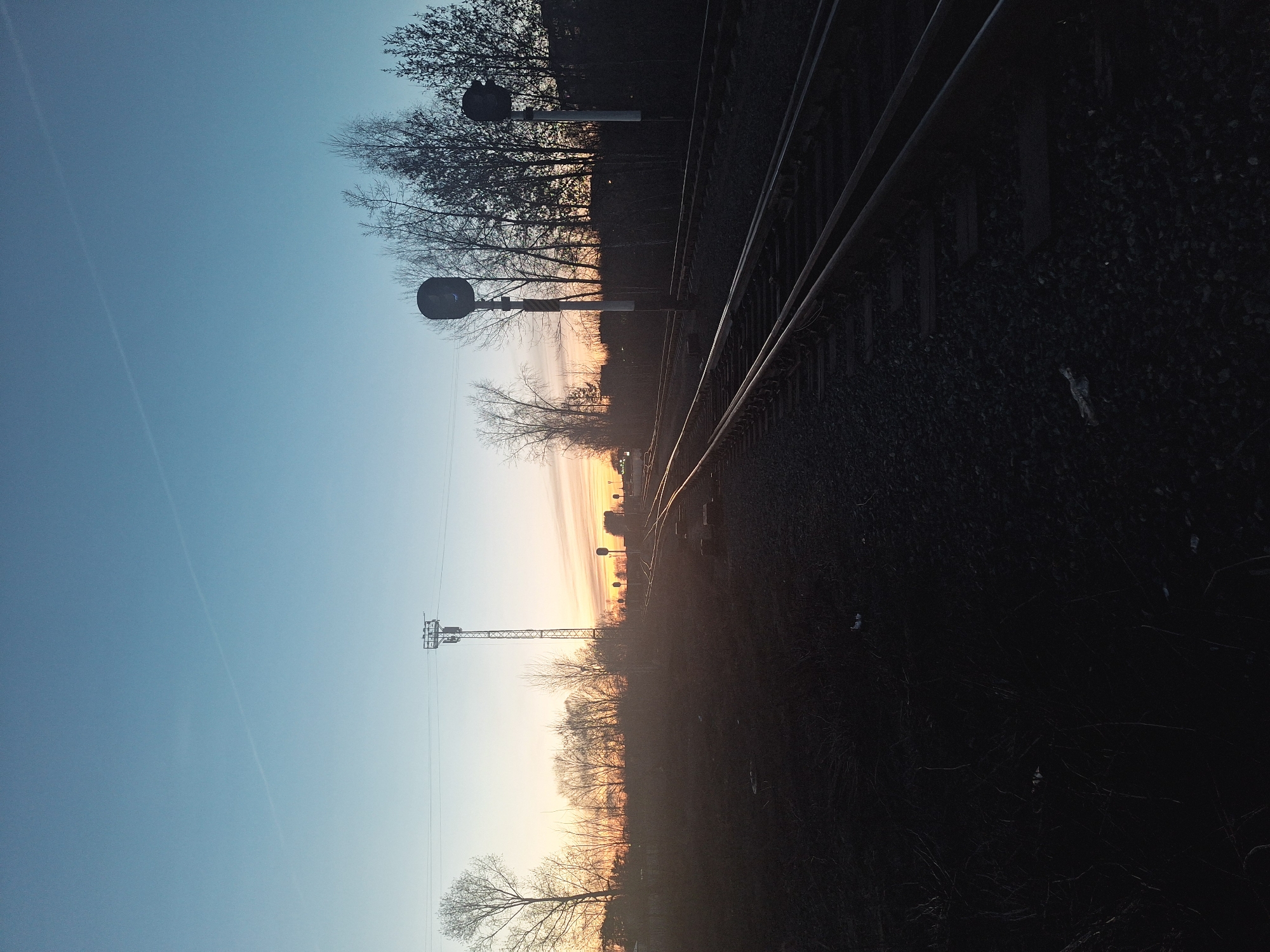 The image depicts a railway track scene at dawn or dusk. The sky transitions from a clear blue to a warm orange and yellow on the horizon. The railway track curves slightly into the distance, bordered by gravel and dark vegetation. Several bare trees and utility poles line the track, creating a silhouette against the sky.
Without additional information, confirming the specific location as Chaikovsky, Russia, is impossible.