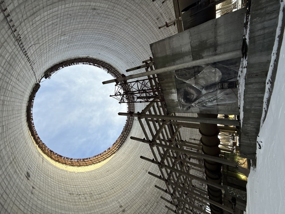 This image presents an upward-looking view from the interior of a massive, cylindrical concrete structure, characteristic of a cooling tower. The scene is set in what is likely the Chernobyl Exclusion Zone, specifically Pripyat, Ukraine.
The vast inner wall of the tower, constructed from light-colored, patterned concrete blocks, curves dramatically upwards, leading to a large circular opening at the top through which a clear blue sky with scattered white clouds is visible. On the right side of the frame, a complex network of concrete beams, metal girders, and wooden scaffolding-like structures ascends vertically, suggesting former industrial infrastructure or construction elements.
A prominent and striking feature is a large, grayscale mural painted on a flat concrete surface integrated into the right-hand structures. The artwork depicts a human face, with a solemn eye, part of a nose, and hands appearing to be covering or holding something close to the face, creating a sense of contemplation or sorrow. Below this mural, several large, dark pipes or conduits descend into the unseen lower parts of the tower.
The ground or base level visible in the bottom right corner is covered in snow, indicating cold weather conditions. Natural daylight illuminates the scene from above, highlighting the immense scale of the structure and the intricate details of the mural. The overall atmosphere evokes a powerful sense of abandonment, industrial decay, and the unexpected presence of human artistic expression within a historically significant and desolate landscape. No visible text is present in the image.