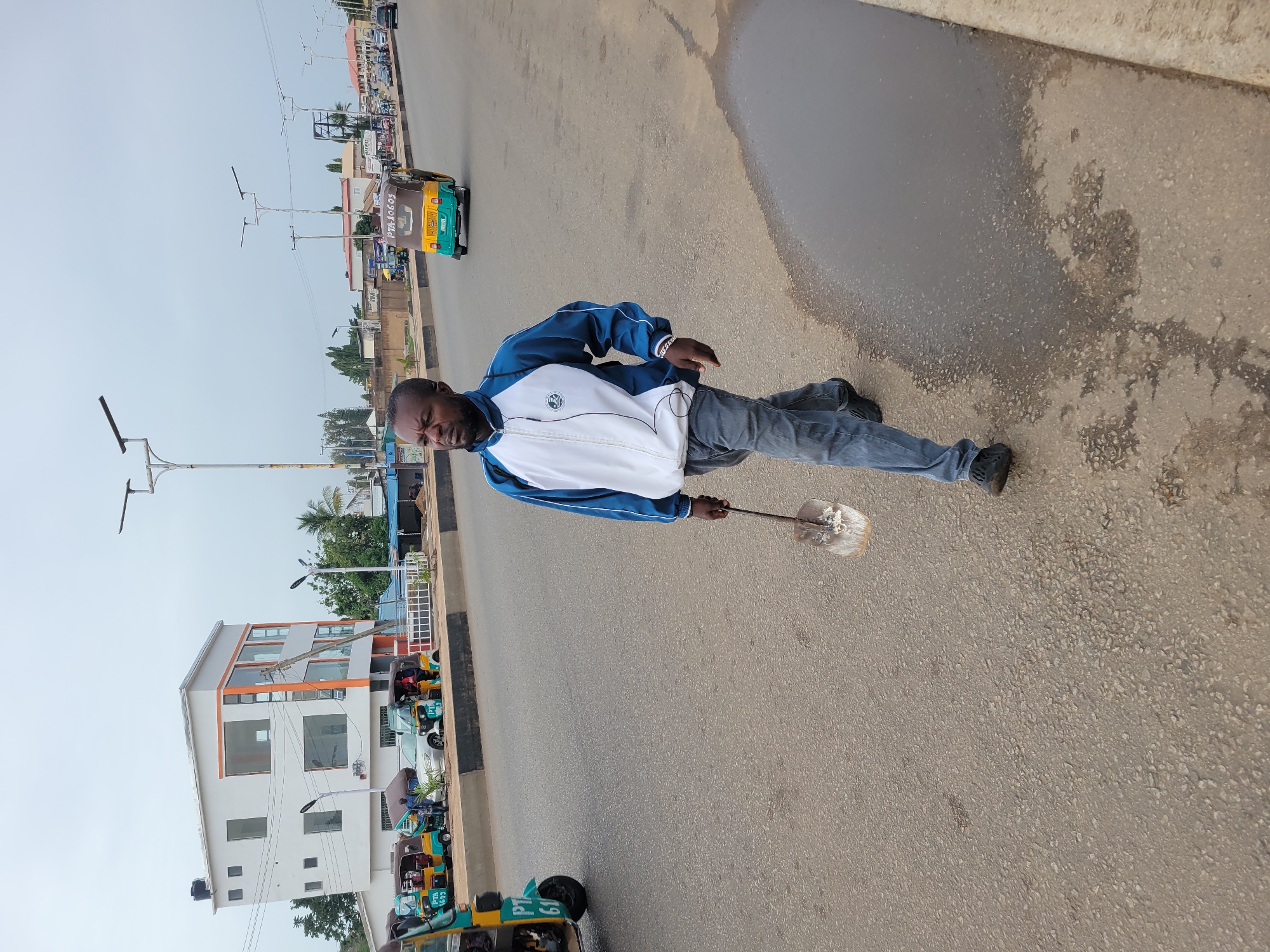 The image captures a man standing in the middle of a road in what appears to be Gura Top, Nigeria. He wears a blue and white jacket and grey jeans, holding a shovel-like tool. The road is paved and shows signs of wear, with some water pooling along the side. In the background, there are buildings, streetlights, and "keke napeps" (auto rickshaws). The overall environment suggests a typical urban scene in a developing country.