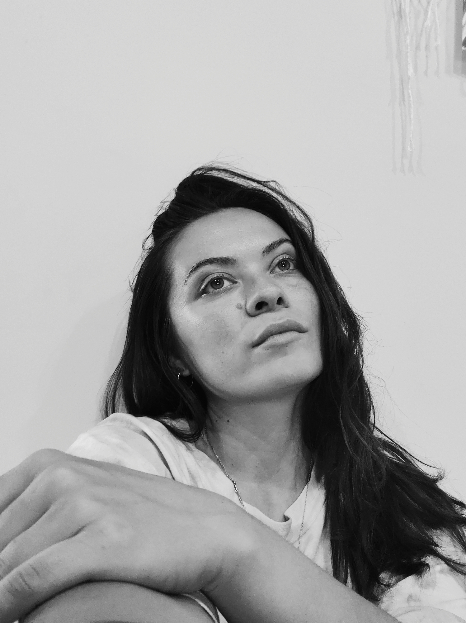 In a black and white, medium shot, a young woman with long, dark, wavy hair is positioned in the lower left quadrant of the frame, looking upwards and to the right. Her left arm is bent and resting across her body, with her left hand visible on her left thigh, which is out of frame. She is wearing a t-shirt and a necklace. Her face is illuminated from the front and slightly from the left, creating subtle shadows. The background is a plain, off-white wall. There are some out-of-focus vertical elements hanging on the wall to the upper right. The scene appears to be indoors, with no indication of a specific city or location.