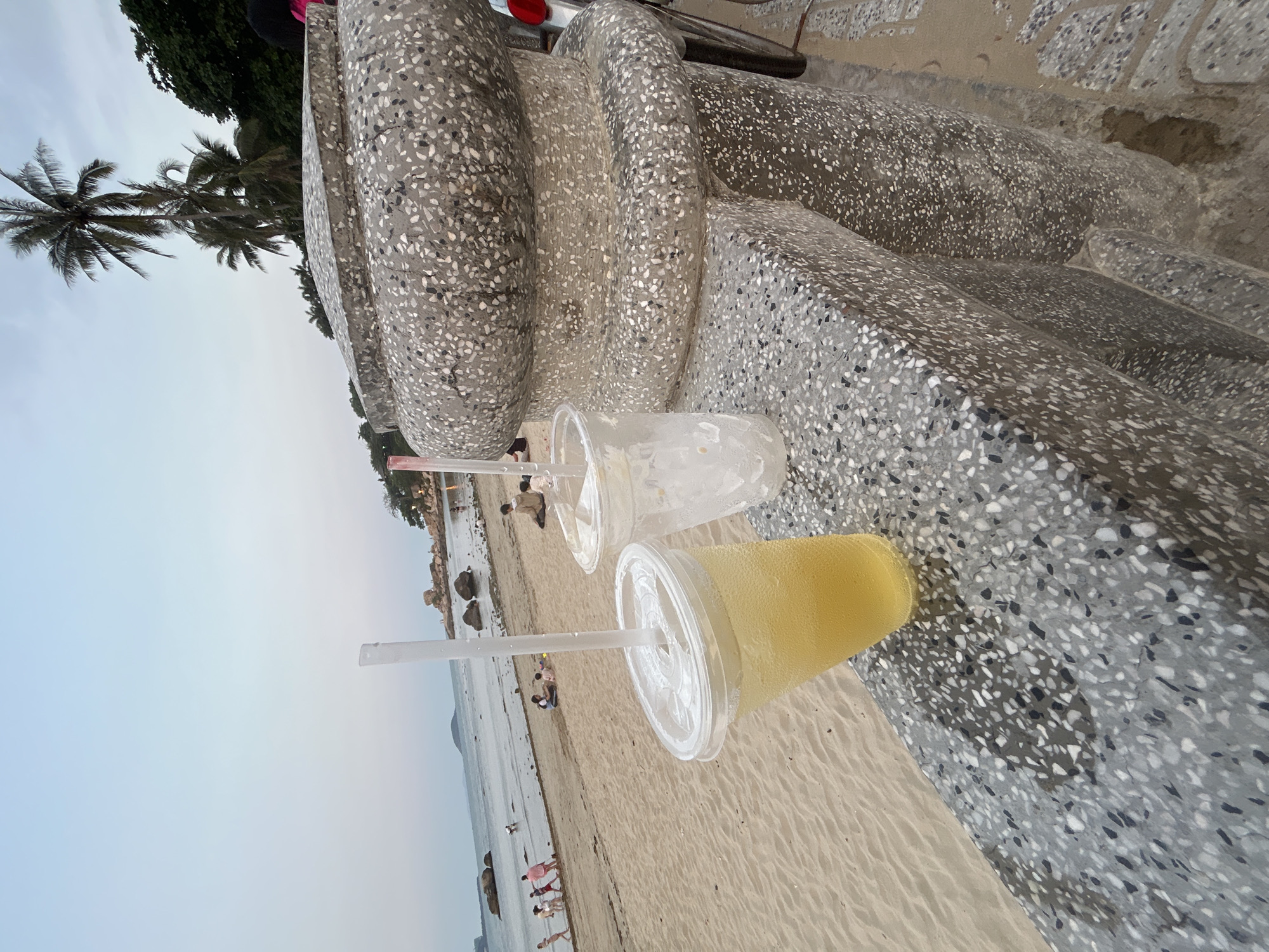 The image shows a beach scene in Bắc Nha Trang, Vietnam. Two plastic cups with lids and straws rest on a concrete structure with a speckled surface, which is likely a public seating area. One cup contains a yellow beverage, and the other appears to be empty with some residue. In the background, a sandy beach stretches along the coastline, dotted with people walking and relaxing. The sea is visible beyond the beach, and the sky is overcast. A palm tree is visible towards the left of the image.
