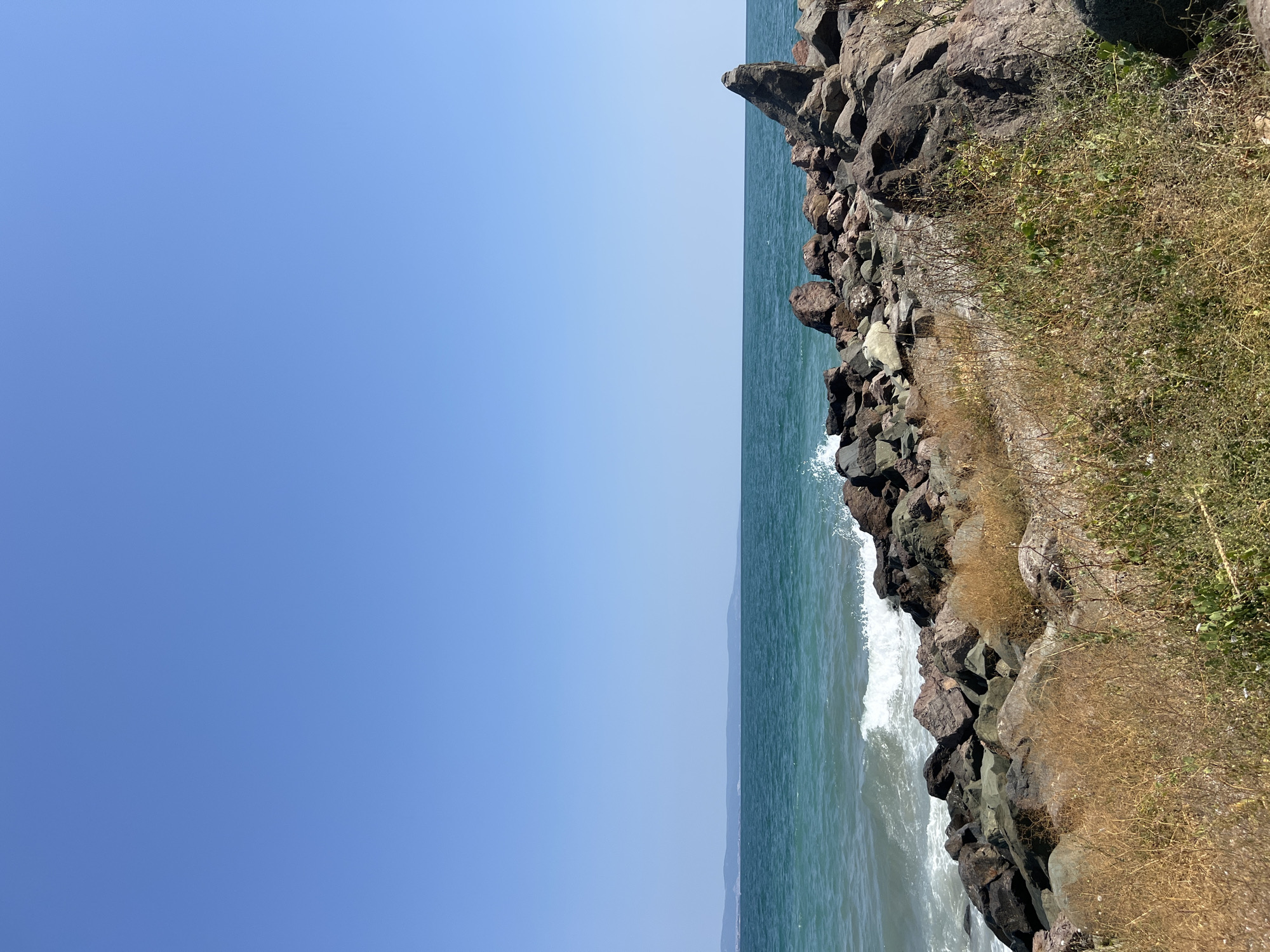 A wide, eye-level shot captures a coastal scene in Pomorie, Bulgaria, on a clear, sunny day with a vibrant blue sky. The turquoise sea stretches to the horizon, where a distant, hazy landmass with rolling hills is visible. Waves crash against a rugged, rocky shore, creating white foam. The foreground is dominated by a pile of large, dark rocks, some appearing reddish-brown and others dark gray, interspersed with dry, yellowed grass and sparse green vegetation. The rocks form a natural breakwater, jutting out into the sea. There are no people or animals visible in the image.