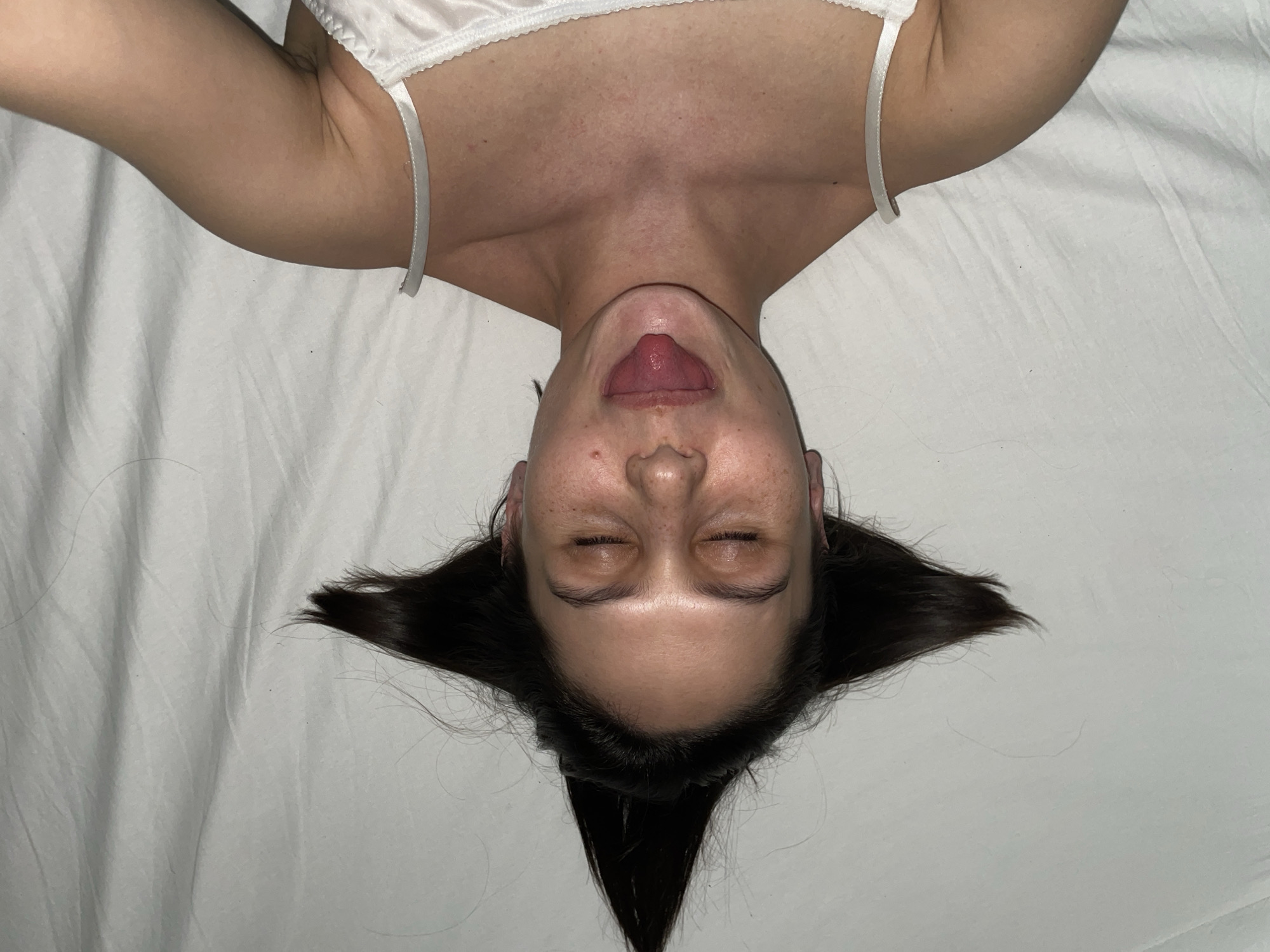 A woman is lying on a white bedsheet with her head at the bottom of the frame. She is wearing a white top with thin straps. Her face is upside down, and her tongue is sticking out. Her eyes are closed, and her hair is spread out on the bedsheet, forming a triangular shape below her head. Her chest and neck are visible above her head. The setting is indoors on a bed, but no location in Izhevsk, Russia can be determined from the image.