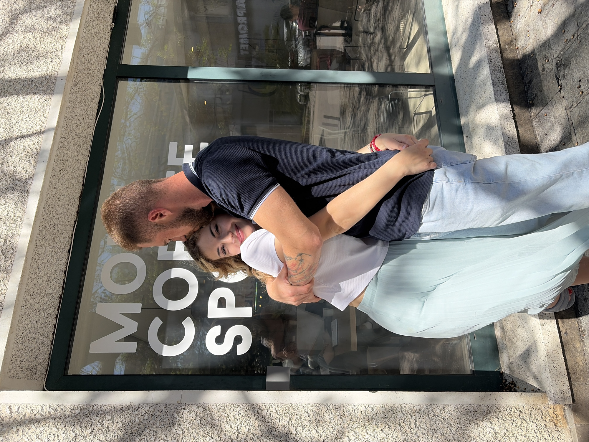 The image depicts a man and a woman embracing affectionately outdoors, likely in Budva, Montenegro. The man, with a light brown beard and short hair, wears a dark blue polo shirt with white trim. He is embracing the woman from behind, resting his head on her shoulder. The woman, with long, light brown hair, is wearing a white t-shirt, a light blue pleated skirt, and light blue jeans. She looks towards the camera with a joyful smile and wears a red beaded bracelet on her left wrist, with a tattoo visible on her right arm.
They are positioned in front of a large glass window of a business, which features the prominent white text "MORE COFFEE SPOT". Reflections in the window reveal interior elements such as tables and chairs, as well as the sky and surrounding trees. The building's facade to the left of the window has a light-colored, textured stucco finish. Strong sunlight illuminates the scene, casting clear shadows and suggesting a bright, clear day, likely late morning or afternoon. The ground beneath them is a paved area. The overall scene conveys a sense of happiness and intimacy.