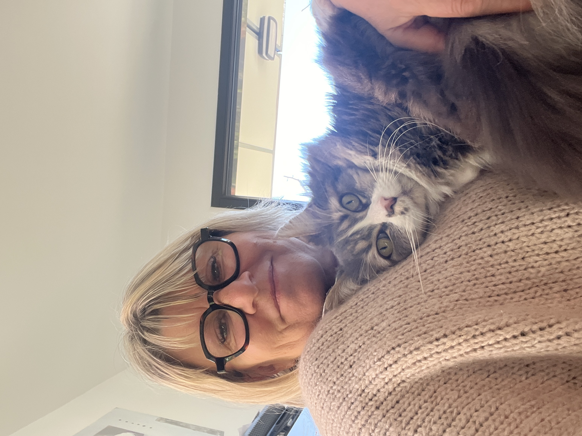 A selfie captured indoors depicts a fair-skinned woman with short, blonde hair and black-framed eyeglasses. She wears a beige knit sweater. A fluffy, gray-and-white cat rests on her right shoulder. The cat's head is visible, displaying its green eyes and long whiskers. A window with a black frame is visible in the upper background, allowing sunlight to filter through. The location of Chambœuf, France is not evident from the provided image.