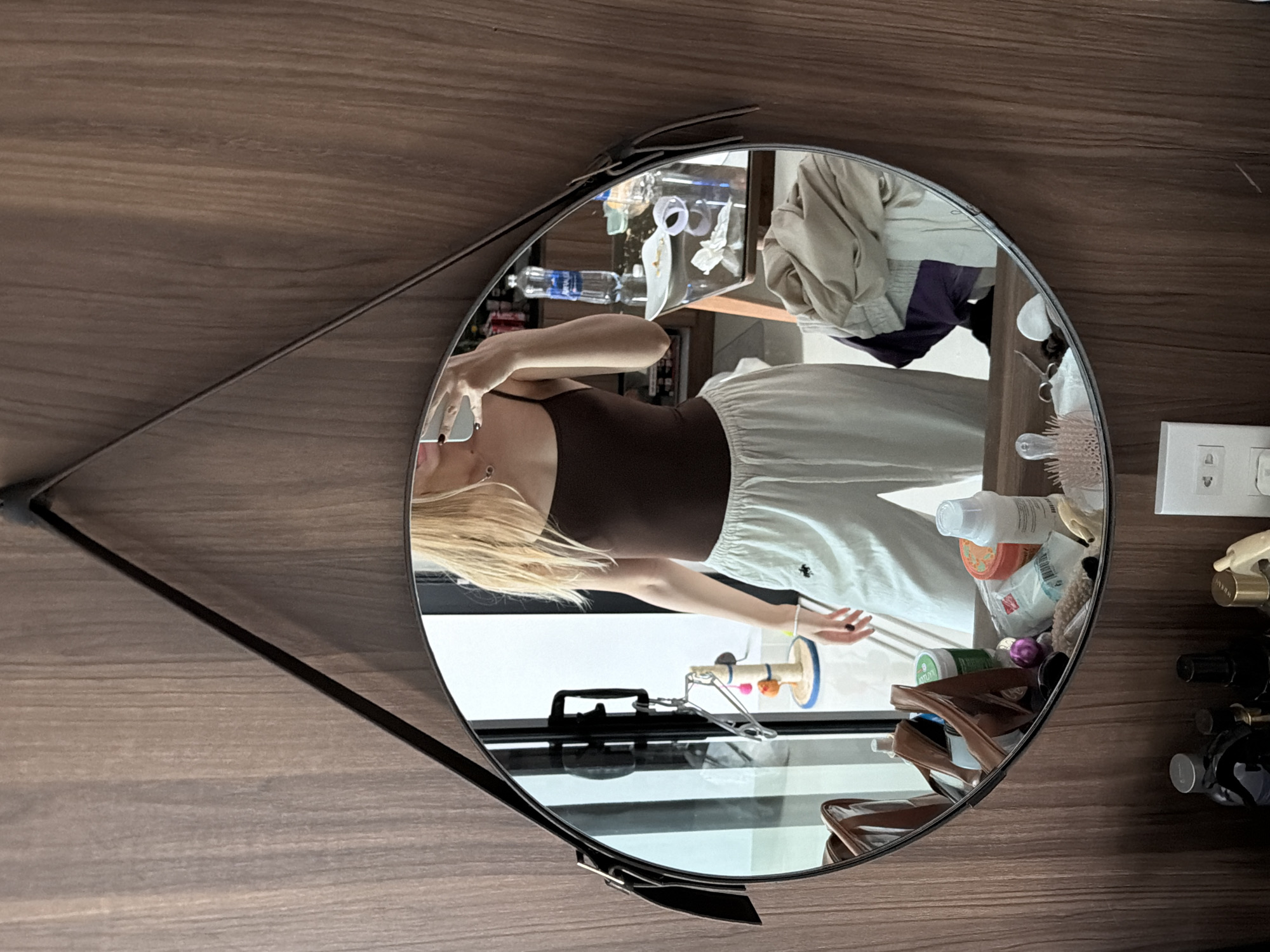 A young woman with blonde hair stands in front of a round mirror, taking a selfie. She is wearing a brown tank top and light-colored pants. The mirror reflects the woman, along with a dresser or table filled with various items. These items include bottles of toiletries, a brown bag, a pink hairbrush, and a water bottle. Behind the woman, a doorway or window is visible, with what appears to be a closet door. The setting is likely an indoor space, possibly a bedroom or dressing area, in Đà Nẵng, Vietnam. The walls are covered in a wood-grain material.