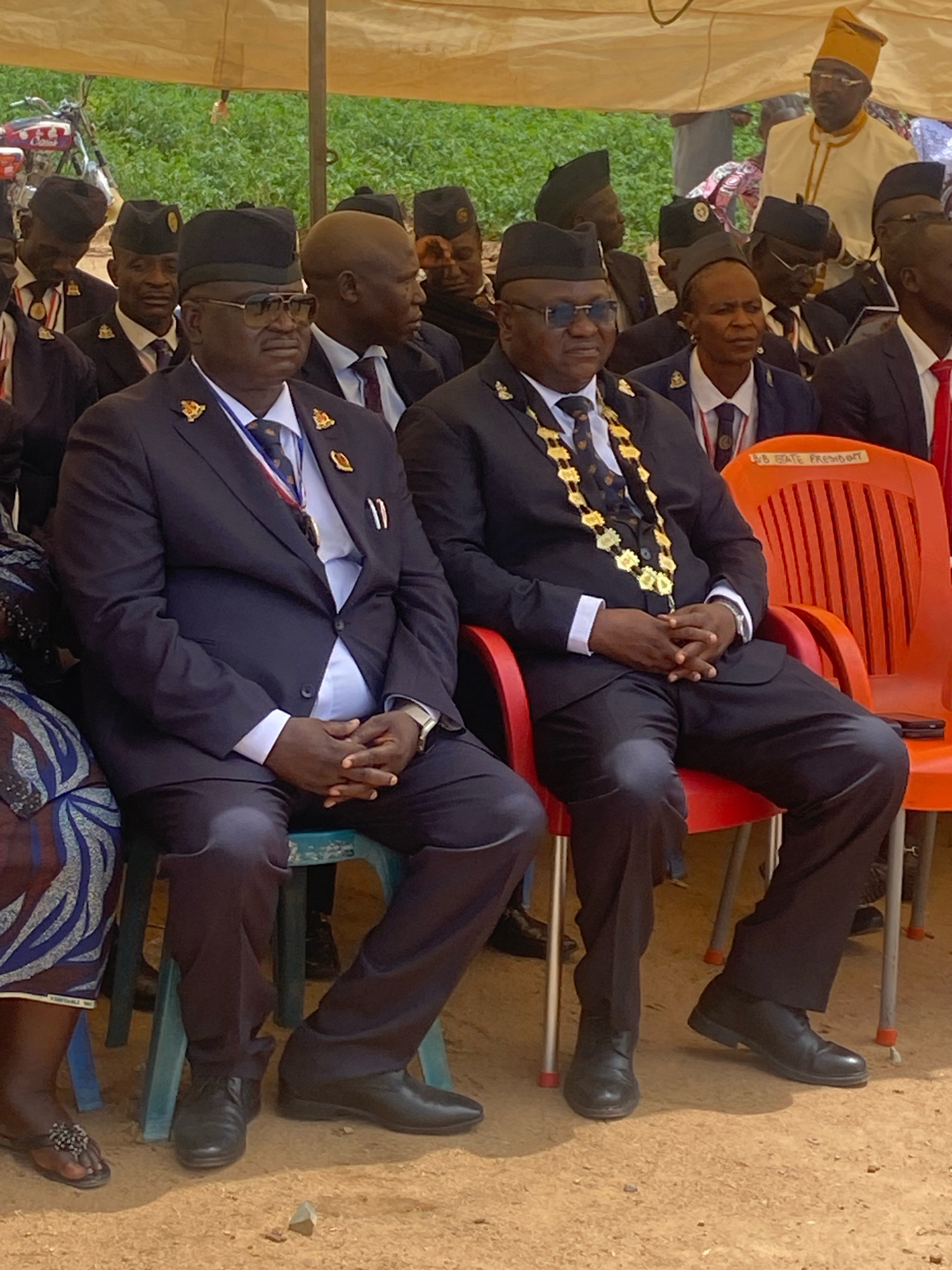 The image depicts an outdoor event, possibly a ceremony or gathering, under a large, beige tent. In the foreground, two men in dark suits and hats are seated on chairs. The man on the left, closer to the camera, wears a black cap with a gold emblem, a dark suit, a light blue shirt, and a tie with a patterned design. He also has a gold chain necklace with circular pendants and a red, white, and blue ribbon draped around his neck. He wears dark-rimmed glasses. The man on the right also wears a dark suit and a black cap with an emblem, a light blue shirt, and a tie. Both men have their hands clasped in their laps. They are seated on teal-colored chairs, with an orange plastic chair visible to the right, bearing a label that reads "SB STATE PRESIDENT."
In the background, several other individuals are visible, some wearing similar attire. To the far left, a person in a patterned fabric is partially visible, seated on a chair. Behind the seated men, a motorcycle with decorations is parked. The ground appears to be dirt. The overall environment suggests a formal gathering in an open, possibly rural, setting. The presence of the label "SB STATE PRESIDENT" on the chair indicates a formal capacity related to a state presidency. The general context of the clothing and decor suggests a significant event.