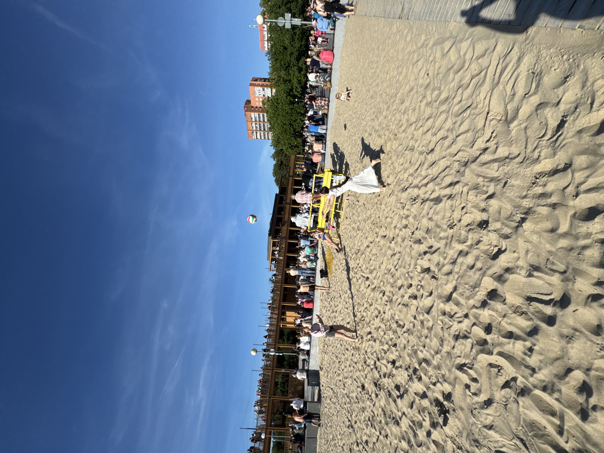 The image depicts a sunny day at a beach in Tallinn, Estonia. In the foreground, sandy terrain with footprints is visible. Several people are scattered across the beach, some playing volleyball. A woman in a white dress is in the process of hitting a volleyball, while another woman in a pink shirt and shorts is also engaged in the game. In the mid-ground, a crowd of people are gathered on a wooden deck, observing the activities on the beach. Behind them, lush green trees and modern buildings, including residential high-rises, can be seen. The sky above is a clear, bright blue, indicating fair weather. The overall scene suggests a lively recreational atmosphere at a beachside location within the city.