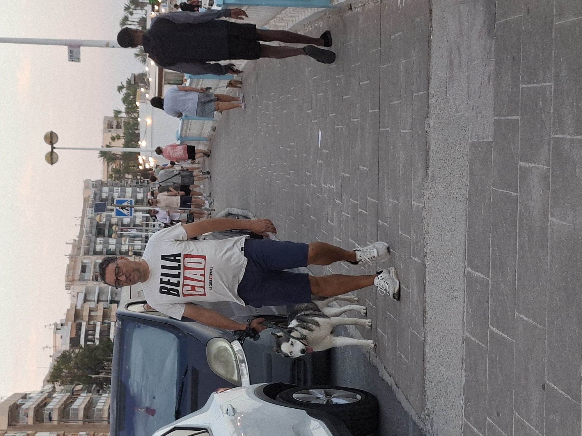 The image depicts a man in his late 40s or early 50s, wearing glasses, a white t-shirt with the text "BELLA CIAO" in red and black lettering, and dark blue shorts. He is standing on a paved walkway, holding a leash attached to a husky dog that is lying on the ground. The dog appears to be a Siberian Husky, with striking blue eyes and a white and grey coat. The man is looking towards the camera with a neutral expression.
Behind the man, a dark blue car is partially visible, as is a white car. The walkway is made of grey paving stones. In the background, there are buildings with multiple stories, characteristic of a coastal urban environment. Palm trees are also visible, suggesting a warm climate. Several other people are walking on the walkway in the background, some appearing to be tourists. The lighting suggests it is either early morning or late evening, likely during the golden hour. The overall scene is one of casual activity on a pleasant day. The text on the man's t-shirt, "BELLA CIAO," is a well-known Italian anti-fascist folk song.