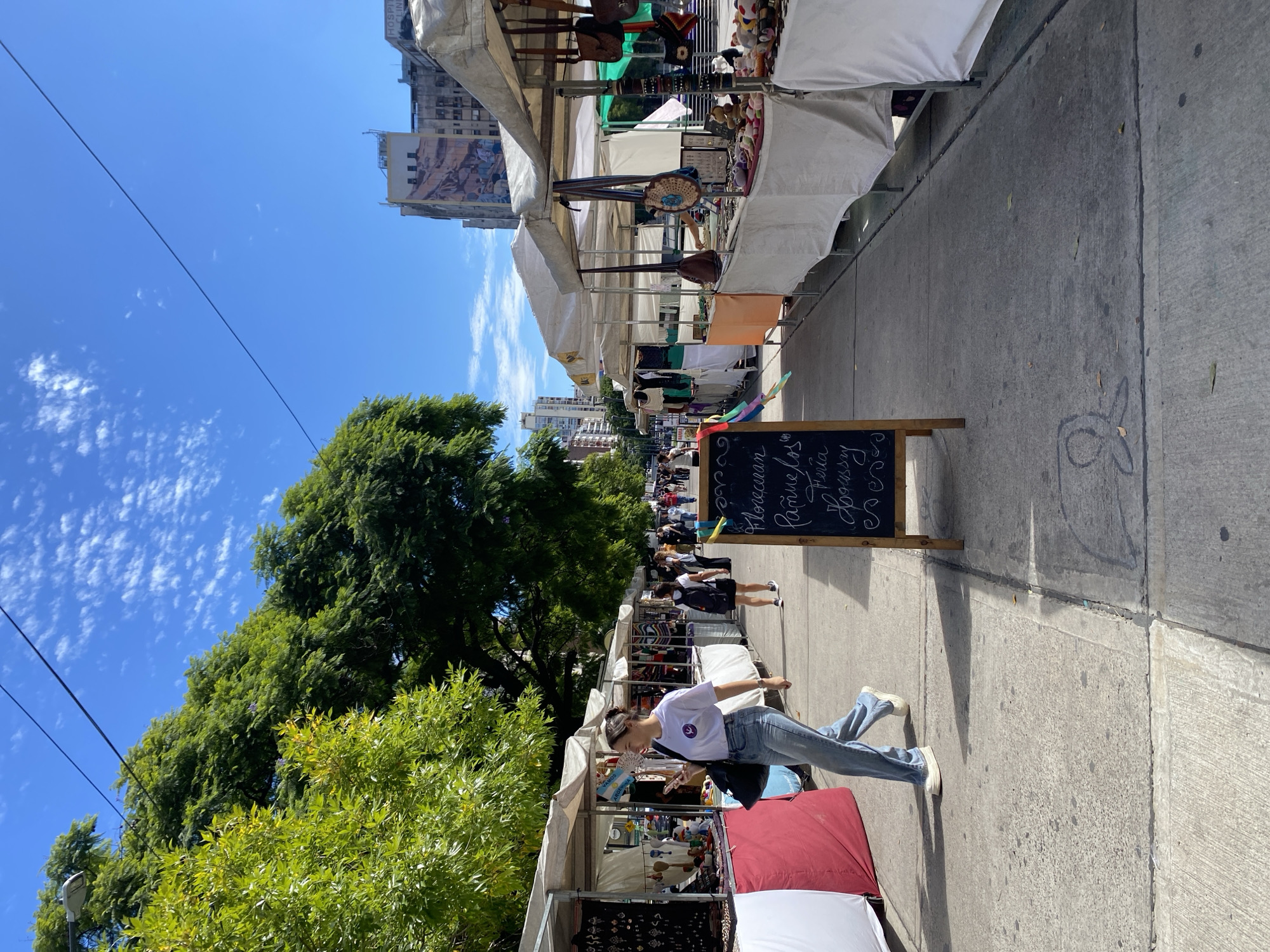 The scene depicts an outdoor street market situated on a wide concrete pedestrian pathway in Plaza Houssay, Balvanera, Buenos Aires, Argentina. The time is daylight, under a clear blue sky with scattered wispy clouds.
Rows of market stalls, each covered by a white canvas awning, line both sides of the pathway, extending into the background. These stalls display various items including jewelry, handmade crafts, bags, and stuffed animals on tables, some adorned with red or orange cloths.
In the left foreground, a young adult female walks from left to right. She has dark hair styled in a bun, wears a white t-shirt with a purple circular logo, light blue wide-leg jeans, and white platform sneakers. She is holding a smartphone in her right hand and carries a dark shoulder bag on her left side.
On the right side of the pathway, in the mid-ground, a wooden A-frame chalkboard sign stands on the concrete. Handwritten white text on the sign includes "Pañuelos," "Feria," and "Houssay."
Further down the pathway, multiple individuals are visible walking and browsing the stalls. One person in a black t-shirt and black shorts, carrying a black backpack, is walking away from the camera on the left side of the pathway.
Large, leafy green trees form a dense canopy along the left side of the market. In the background, multi-story urban buildings are visible, including the Hospital de Clínicas José de San Martín, identifiable by a large mural on its facade and partial signage reading "HOSPITAL CLINI." Overhead, dark utility wires traverse the sky. The concrete pavement features visible expansion joints and minor surface marks.