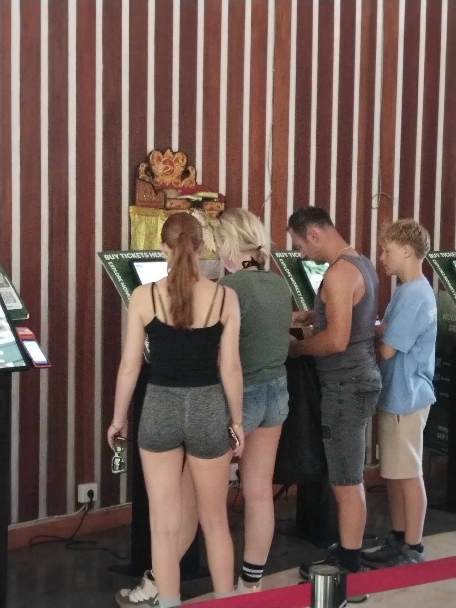 In Ubud District, Indonesia, a group of four individuals, two women and two males, stand before ticket vending machines. The women, one with blonde hair and the other with auburn hair in a ponytail, are positioned to the left of the machines. The auburn-haired woman, wearing a black tank top and grey shorts, holds a phone in her left hand. The blonde-haired woman, dressed in a green top and denim shorts, stands beside her. To the right, a man in a grey tank top and dark shorts interacts with a machine, while a younger male in a blue t-shirt and light-colored shorts stands next to him, facing the machines. The background consists of a wooden wall with vertical brown and white stripes and a decorative golden object on a pedestal. The floor is dark, and a red barrier tape is visible in the foreground.