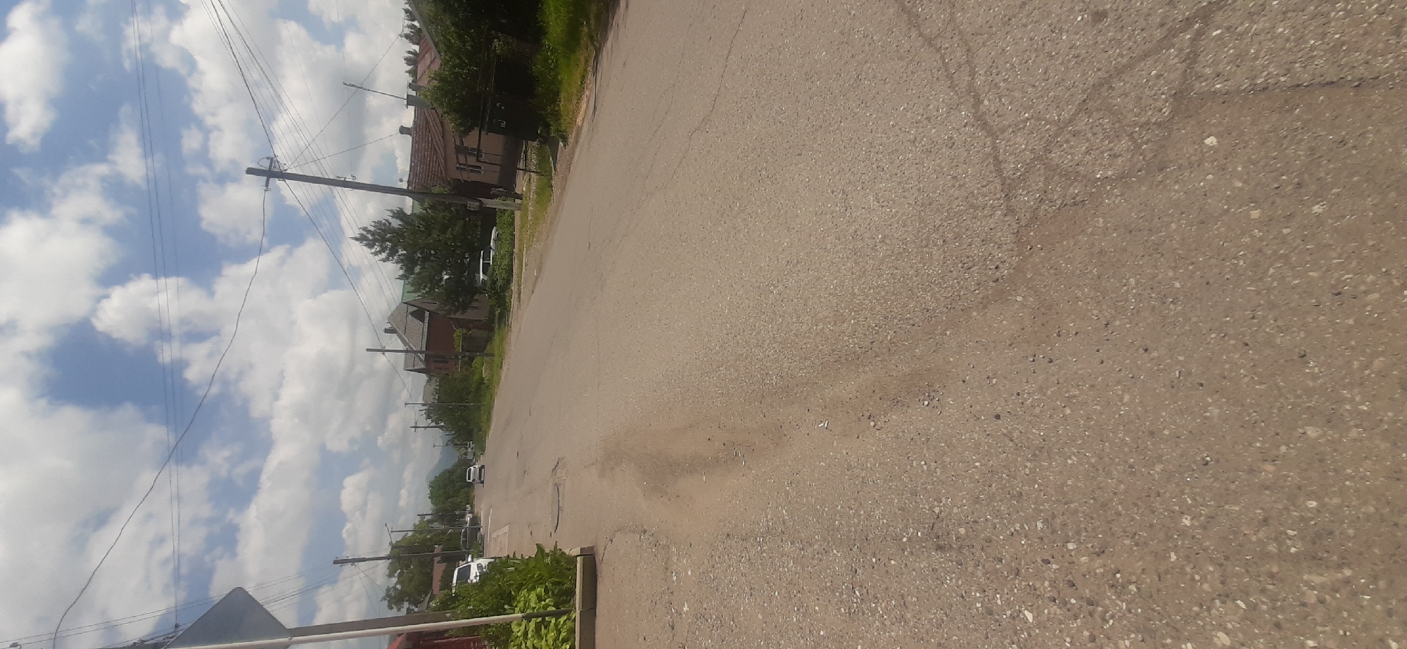 A road scene in Yessentukskaya, Russia, features a grey asphalt road with visible cracks and gravel on the sides. Flanking the road are houses with tiled roofs, some featuring red brick. Dense green trees and bushes line the sides of the road, with a clear blue sky dotted with white clouds overhead. A few cars are visible on the road and parked on the side. Telephone poles and wires stretch across the scene. The time of day appears to be daytime, with the sun illuminating the environment. The overall impression is of a quiet, residential street in a rural or semi-rural setting.