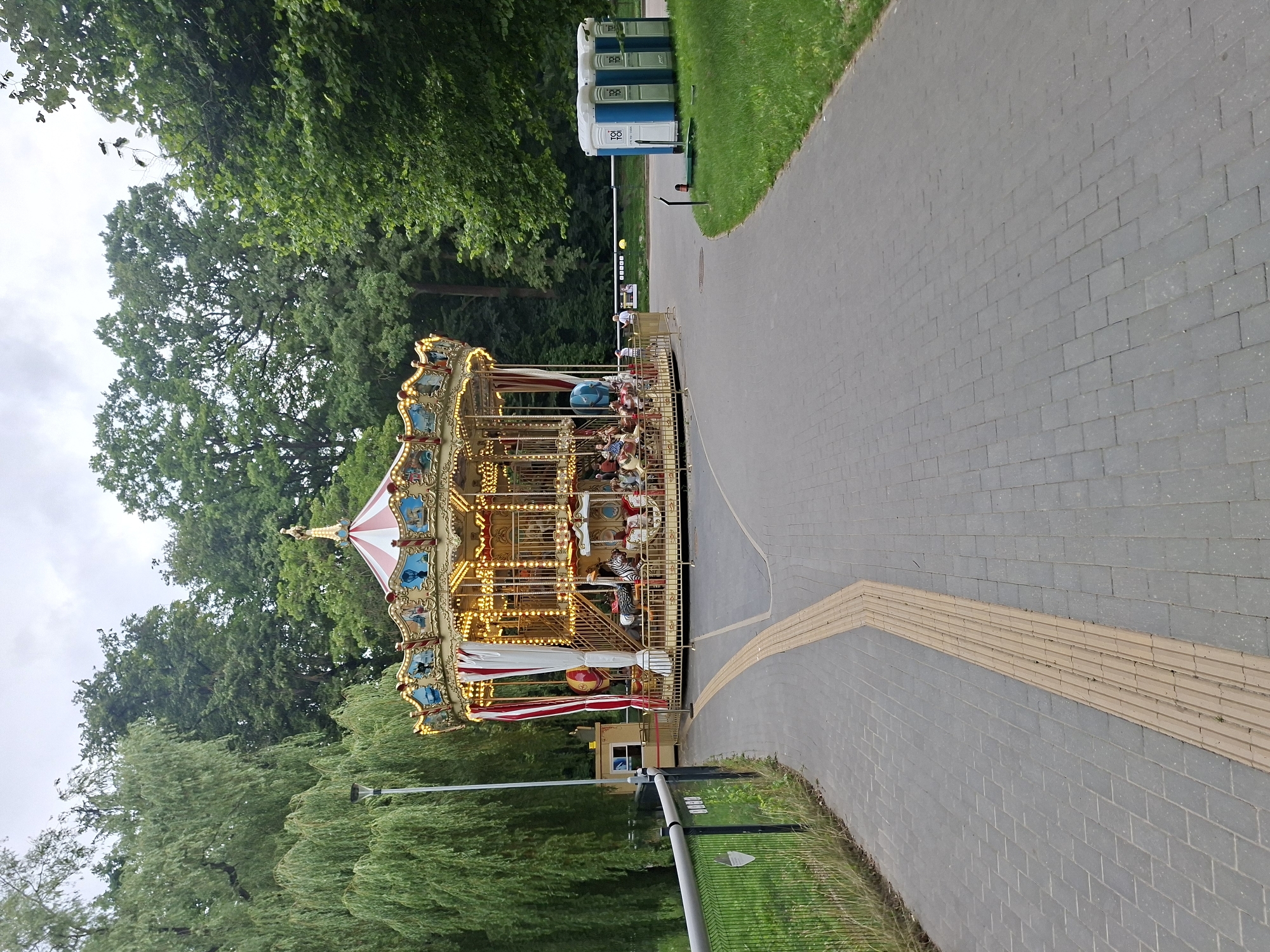 The image shows a carousel in a park on a cloudy day. The carousel is brightly lit and decorated with red and white curtains. Several people are riding on the carousel, which has various animal figures. To the left of the carousel, there is a pathway made of gray pavers with a tactile paving strip for the visually impaired. A metal fence separates the pathway from a grassy area with trees. To the right of the carousel, there is a grassy area with three portable toilets. In the background, there is a dense line of trees. The image was taken in Kaunas, Lithuania.