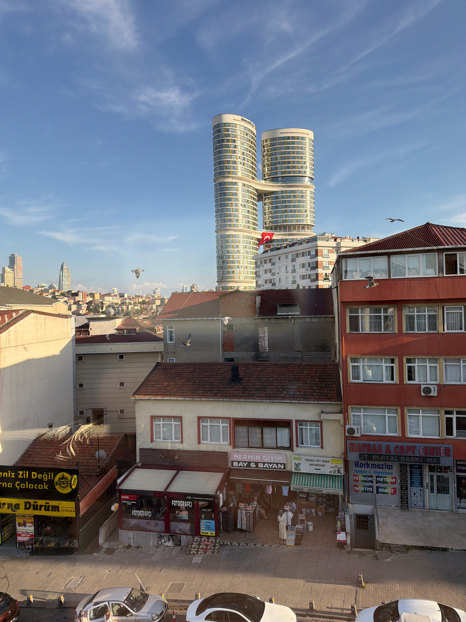 The image depicts a street scene in Ataşehir, Turkey, with a clear blue sky featuring scattered clouds and two distinctive, modern, curved high-rise buildings in the background. In the mid-ground, there is a cluster of older, multi-story buildings. The foreground shows the street level with several shops. One shop has a yellow sign with black text and an image of a flame, followed by shops with signs indicating "FOTOGRAFÇI" (photographer) and "Kodak". Another shop has a red sign with white text that reads "MATBAA & COPY CENTER" and "Korkmazlar Matbaa". A Turkish flag is visible on one of the buildings. Several white birds, likely seagulls, are flying in the sky. Cars are parked on the street in the foreground, and some people are visible on the sidewalk in front of the shops. The overall scene suggests a busy urban environment with a mix of modern and older architecture.