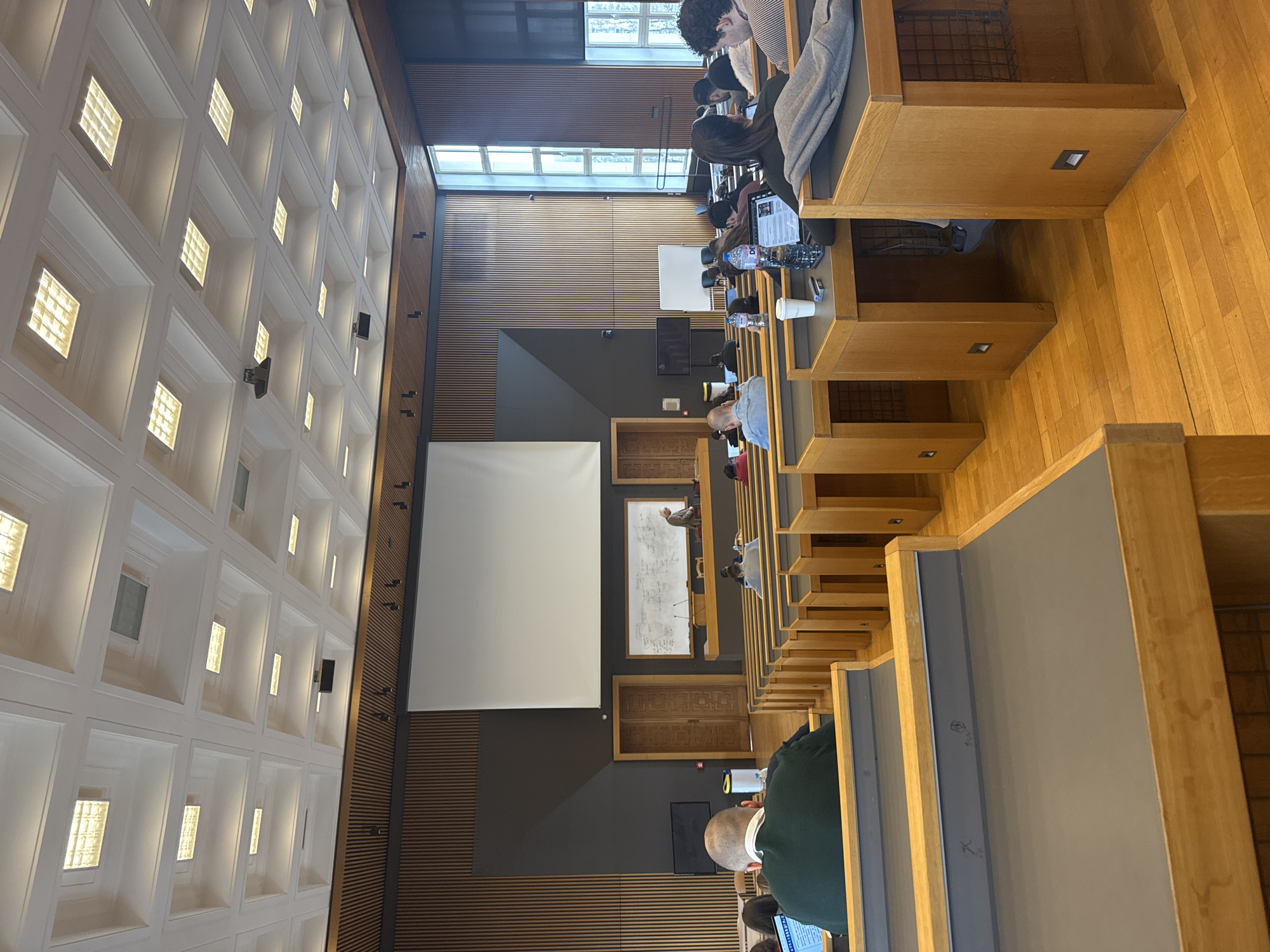 The image depicts an interior view of a lecture hall in Sofia, Bulgaria, characterized by tiered wooden seating arrangements occupied by students. The room features a high, coffered ceiling with integrated lighting fixtures. A large white screen hangs on the wall to the left. A smaller whiteboard with writing on it is positioned behind and above some of the students, with a wooden door below. Natural light enters through windows located high on the wall, with vertical wood paneling below. Students are seated at desks with gray tops, some using laptops or taking notes. The setting appears to be a university lecture in progress.
