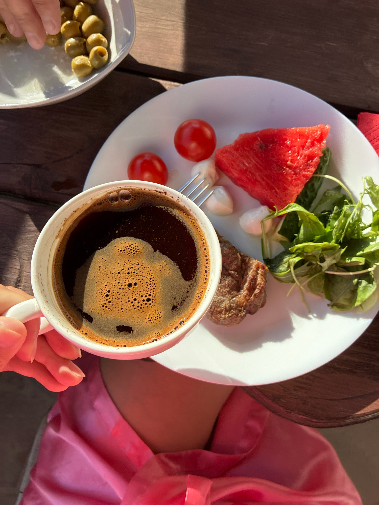 A high-angle close-up shot captures a breakfast spread on a wooden table in Abrau-Dyurso, Russia. A white mug of dark coffee with foamy crema is held by a person's hand in the lower left corner. On a white plate to the right, there is a slice of watermelon, two cherry tomatoes, mozzarella balls, a piece of grilled meat, and a small green salad. In the upper left corner, a white dish contains a pile of green olives, with a hand reaching for them. The scene is illuminated by bright sunlight, casting shadows on the table. The person is wearing a pink silk garment.