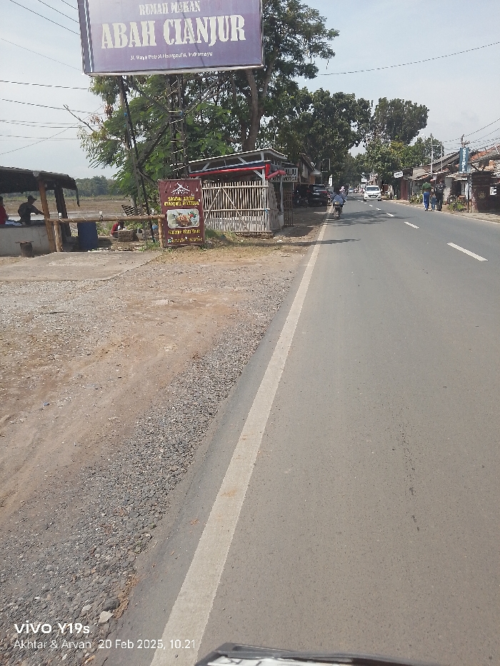 This image captures a daytime scene along a road in Patrol, Indonesia. The setting appears to be a rural or semi-urban area, with commercial establishments lining one side of the road and what looks like open land or fields on the other.
In the foreground, a dusty gravel shoulder meets a paved asphalt road marked with a white lane line. On the left, several people are visible sitting under a simple, open-air shelter with a wooden frame and a thatched or corrugated roof, possibly dining or resting. Beyond this, a large blue billboard prominently displays "RUMAH MAKAN ABAH CIANJUR" (Abah Cianjur Restaurant) and an address: "Jl. Raya Patrol Haurgeulis, Indramayu." Below this sign, a smaller reddish-brown sign advertises "SATE AYAM / MANDUL PATTYUA," showing an image of food.
Further down the road on the right, various buildings and storefronts are visible. People are walking along the roadside, and a person on a motorcycle is driving away from the viewer. Cars are also present further down the street. The weather appears sunny with clear skies, indicated by the bright, even lighting.
The visible text also includes a partial sign on a building to the right of the main billboard that reads "CALJU MOTOR." A watermark at the bottom left corner states "VIVO Y19s Akhtar & Arvan 20 Feb 2025, 10.21," indicating the device used and the time and date the photo was taken.