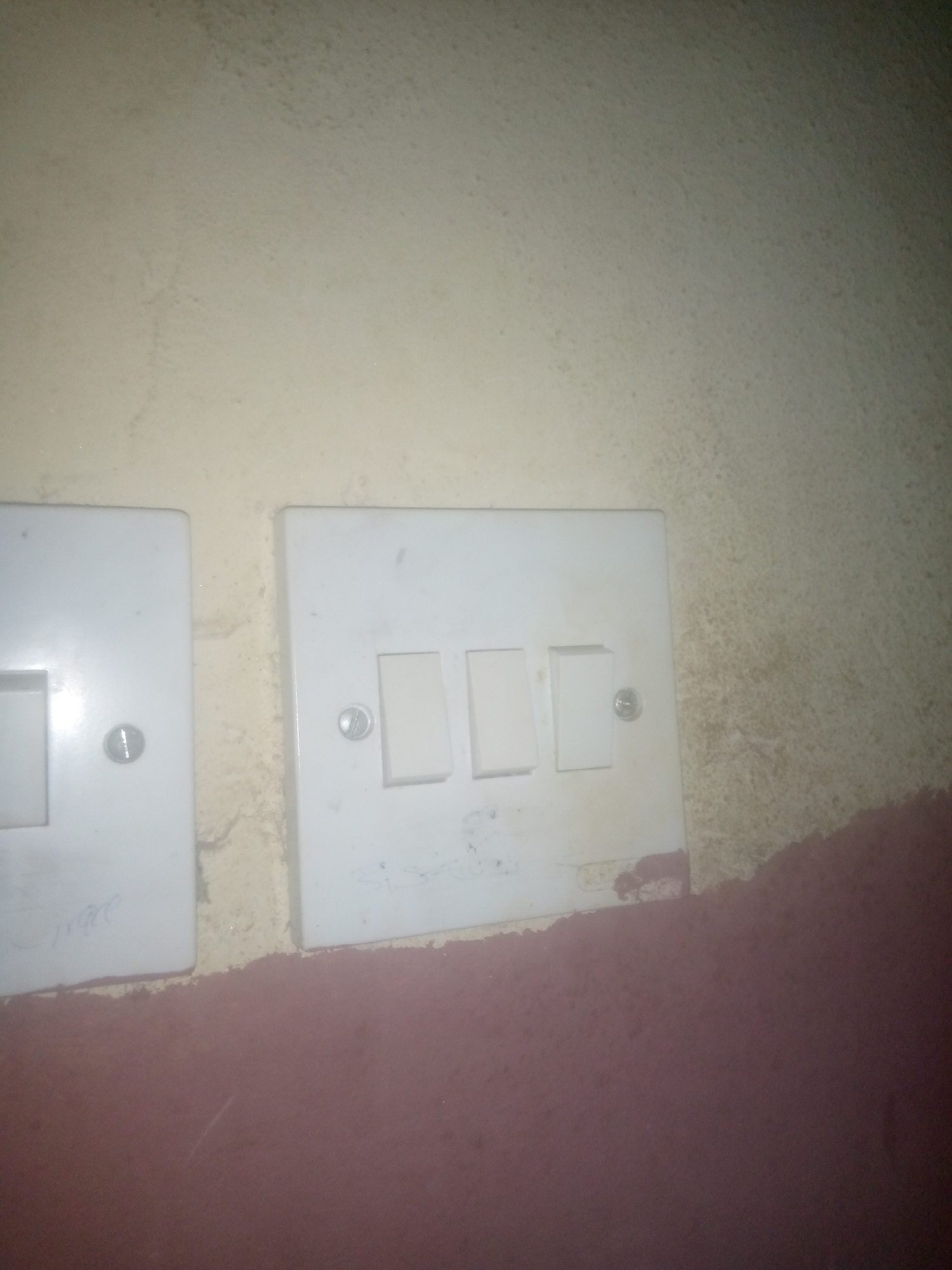 The image shows two white electrical switch plates mounted on a textured, light-colored wall. The switch plate on the right has three toggle switches, two of which are visible and appear to be in the down position. The switch plate on the left has a single toggle switch, also visible and in the down position. A silver screw is visible in the center of each switch plate. The wall below the switch plates is painted a dark reddish-brown color. There are some smudges and discoloration on the wall and the switch plates, suggesting age or dirt. The lighting is dim, casting shadows. The scene appears to be indoors, likely in a residential or commercial building.