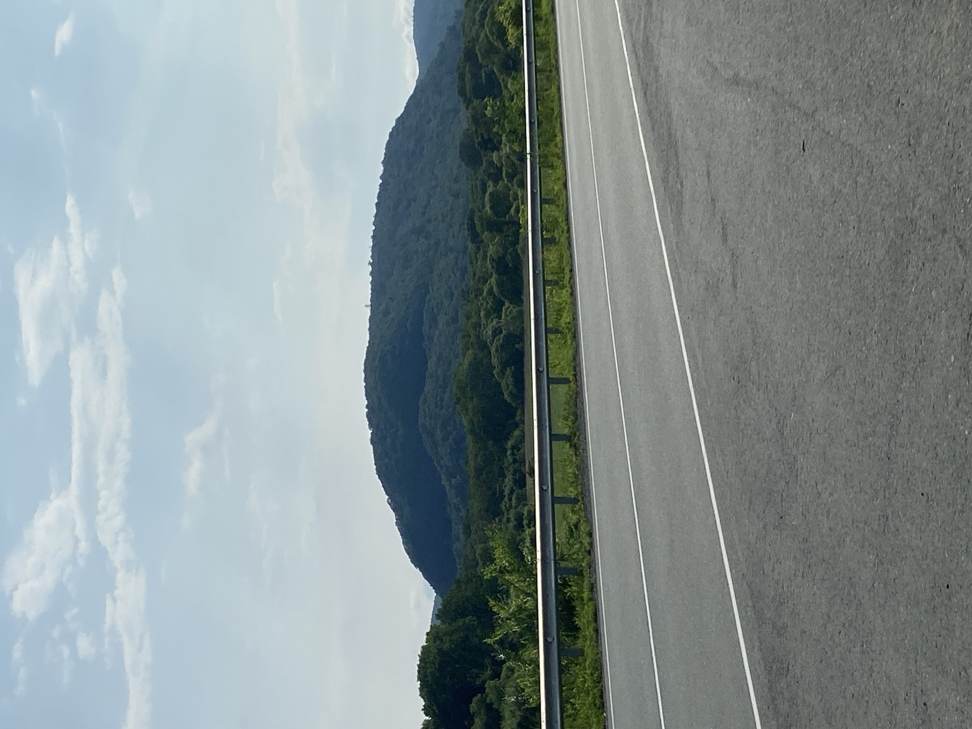The image shows a view from a roadside towards a large, forested hill under a partly cloudy sky. A grey asphalt road with white lane markings occupies the foreground. A metal guardrail runs horizontally across the middle ground, separating the road from a grassy area and a dense line of green trees. Beyond the trees, the main subject is a domed hill covered in dark green foliage. The sky above is a pale blue with scattered white clouds. The scene appears to be located in a rural or natural environment, possibly on the outskirts of a town or city like Kravtsovka, Russia. No people or animals are visible in the image.