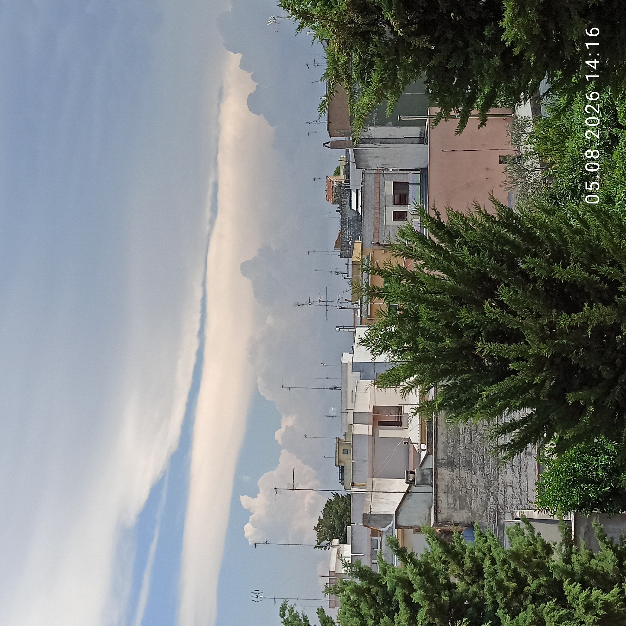 The image depicts a rooftop view of buildings in Bitonto, Italy, under a cloudy sky. The foreground is dominated by two evergreen trees on either side, with their branches partially obscuring the view of the rooftops. The buildings are a mix of styles and ages, with white and beige facades, terracotta tile roofs, and some featuring stone elements. Numerous antennas and satellite dishes are visible on the rooftops, indicating a residential or commercial area. The sky is filled with large, dramatic clouds, some appearing white and puffy, others darker and more ominous, suggesting potential weather changes. In the bottom right corner of the image, a timestamp reads "05.08.2026 14:16". There are no people or discernible actions taking place in the scene.
