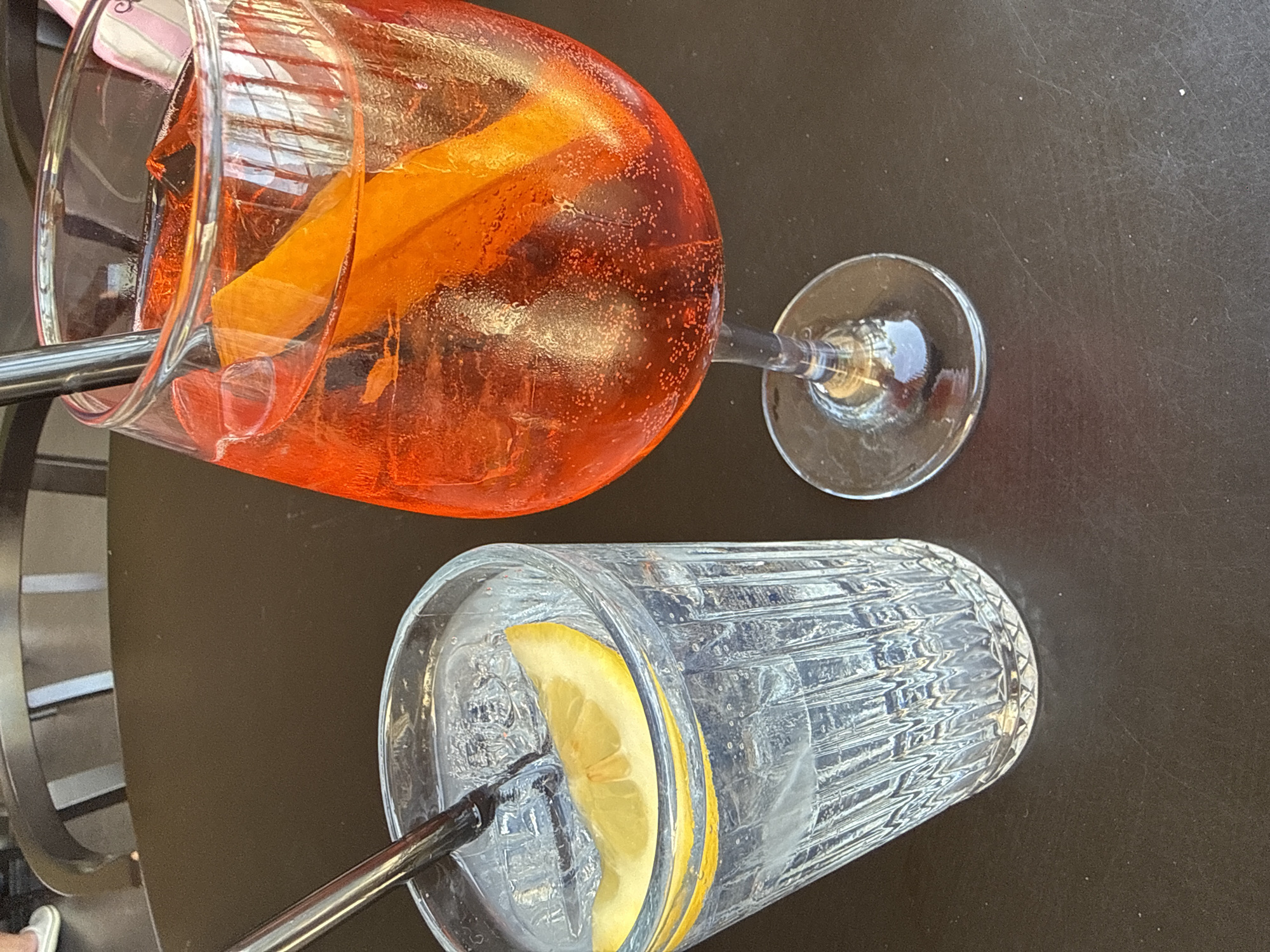 Two drinks are on a dark table, one is a clear drink with ice and a lemon slice in a fluted glass with a straw, and the other is an orange drink with ice and a slice of orange in a wine glass with a straw. This image appears to be taken at a cafe in Bodrum, Turkey, as indicated by the light reflecting off the drinks, suggesting an outdoor setting with bright sunlight characteristic of the region. The drinks are positioned on a table, with hints of chairs and possibly other patrons visible in the background, suggesting a social or dining environment. The focus is on the drinks, highlighting their refreshment and vibrant colors.