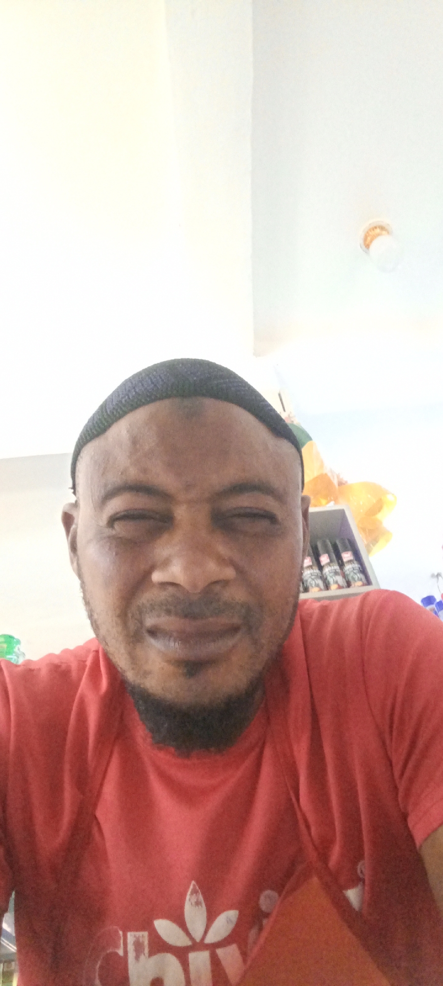 A man with dark skin and a black kufi cap is facing the camera. He is wearing a red t-shirt with a white design. He has a short beard and his eyes are partially closed. In the background, there is a white ceiling with a light fixture and a shelf with objects, including yellow plastic bags and what appear to be spray cans. The location is Fagge D2, Nigeria.