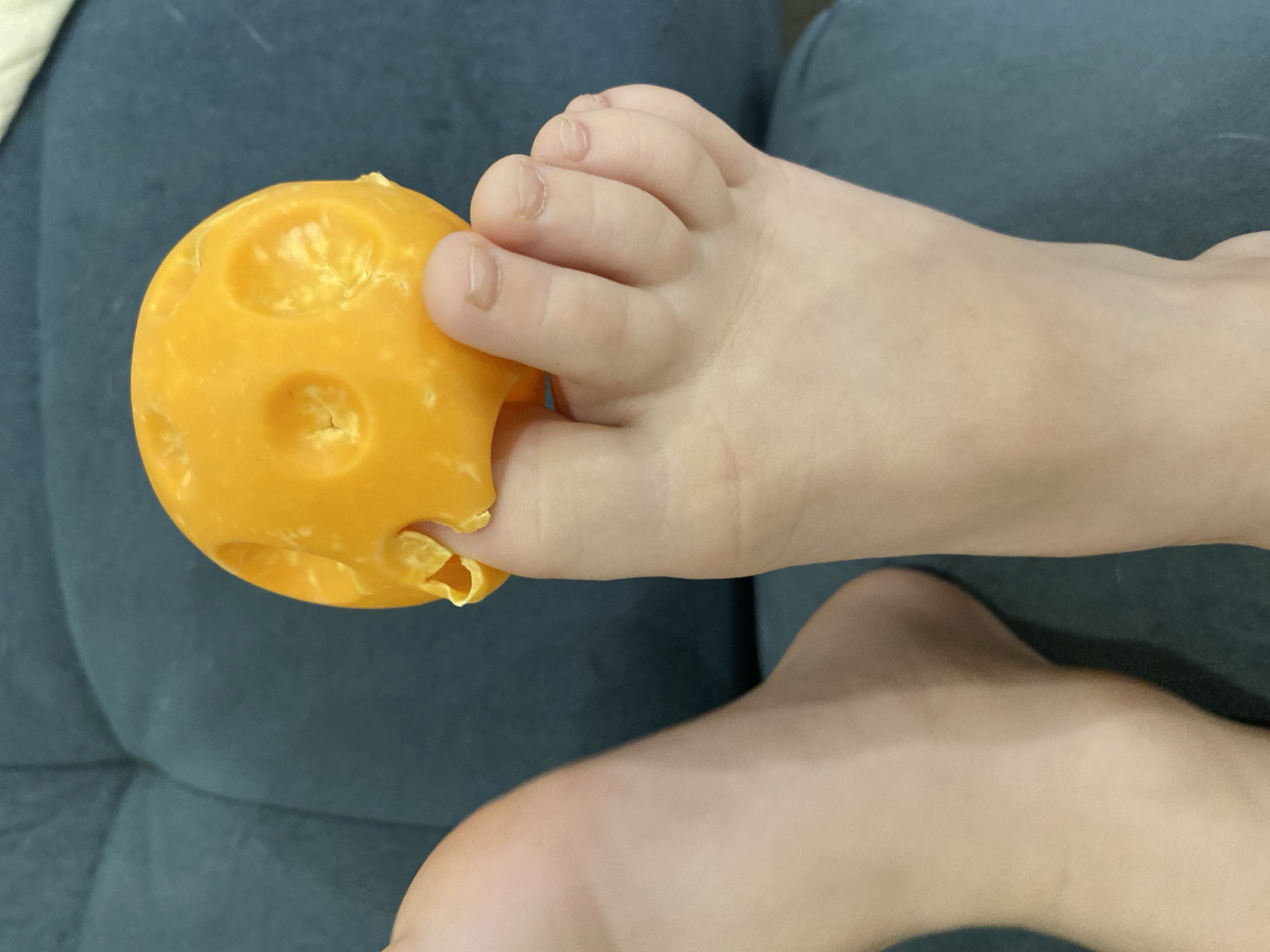 The scene depicts a close-up view of two human feet in what appears to be an indoor, domestic setting in Chaikovsky, Russia. The right foot, positioned centrally, is actively gripping a bright orange, spherical object between its toes. The object features multiple circular indentations and irregular cut-outs, suggesting a textured or damaged surface. The left foot is partially visible on the left side of the frame. Both feet are bare, and their toenails are short and unpainted. The immediate background consists of a dark teal upholstered surface with visible stitching, consistent with furniture like a sofa or armchair.