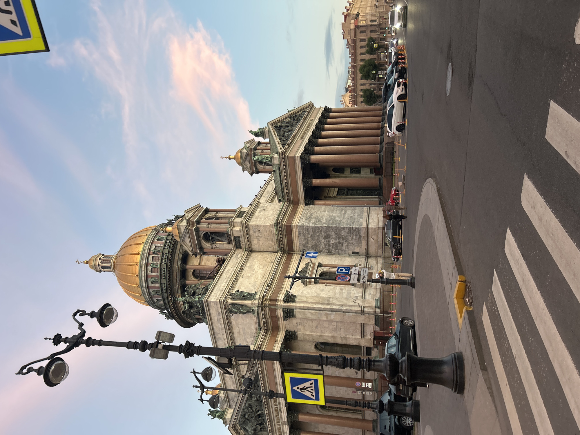 An exterior, eye-level, medium shot captures a golden-domed building with classical architecture, possibly St. Isaac's Cathedral in Saint Petersburg, Russia, under a partly cloudy sky with pink and blue hues. The building features large columns, sculptures, and intricate detailing. In the foreground, a street with a pedestrian crossing is visible, along with parked cars and streetlights. A few people are present on the sidewalk and crossing the street. The overall impression is one of urban grandeur and architectural significance.
