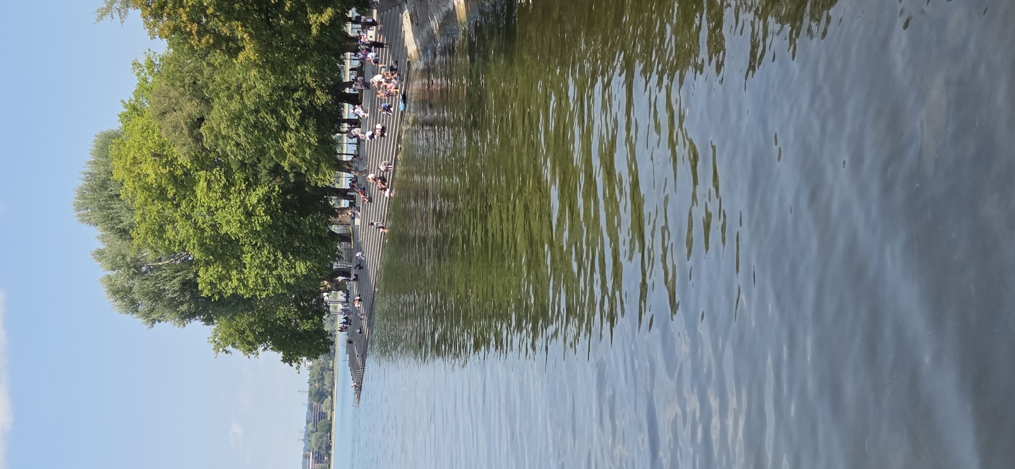 The image depicts the waterfront of Bregenz, Austria, specifically the area near the Festspielhaus. In the foreground, calm, rippling water reflects the greenery of the trees and the blue sky with scattered clouds. The water is a murky green, suggesting depth or sediment.
Along the shore, a wide, stone-tiered embankment serves as seating for numerous people. These individuals are engaged in various leisure activities: some are sitting and conversing, others are lying down, and a few are wading in the shallow water at the edge. There are several beach towels and bags scattered on the steps.
Behind the embankment, a row of mature, leafy trees provides shade, their dense foliage a vibrant green against the bright sky. In the distance, across the water, the cityscape of Bregenz is visible, characterized by buildings and some industrial elements, including power lines. The overall scene conveys a sense of a warm, sunny day, with residents and visitors enjoying the recreational amenities of the lakefront.