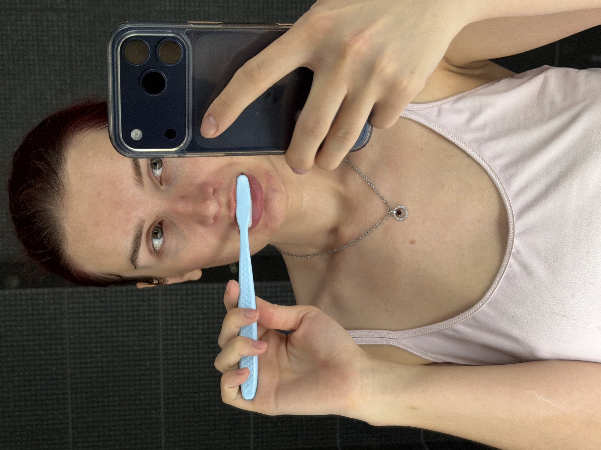 A person is taking a selfie while holding a blue toothbrush near their mouth. They are also holding a smartphone with a clear case. The person has red hair and is wearing a light pink tank top and a silver necklace. They appear to be in a bathroom with dark grey tiles in the background. The location is unknown and there is no evidence indicating that the photo was taken in Chaikovsky, Russia.