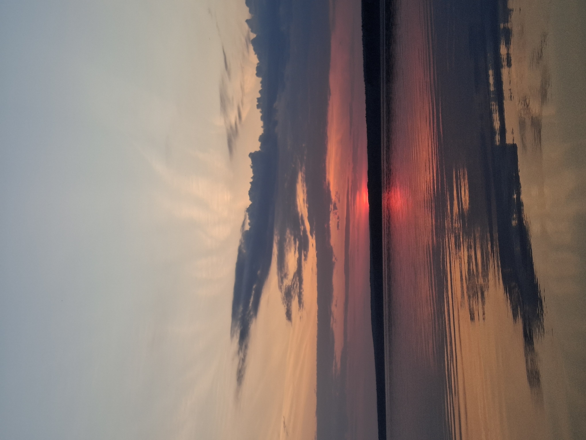 The image captures a sunset reflected in water, likely a lake or river. The sky exhibits hues of orange, pink, and gray, with dark clouds along the horizon. A vertical dark line on the right side suggests a shoreline or landmass. The water reflects the colors of the sky and clouds, with visible ripples. The image is likely from Chaikovsky, Russia, based on the prompt. No people, specific objects, or interactions are discernible.